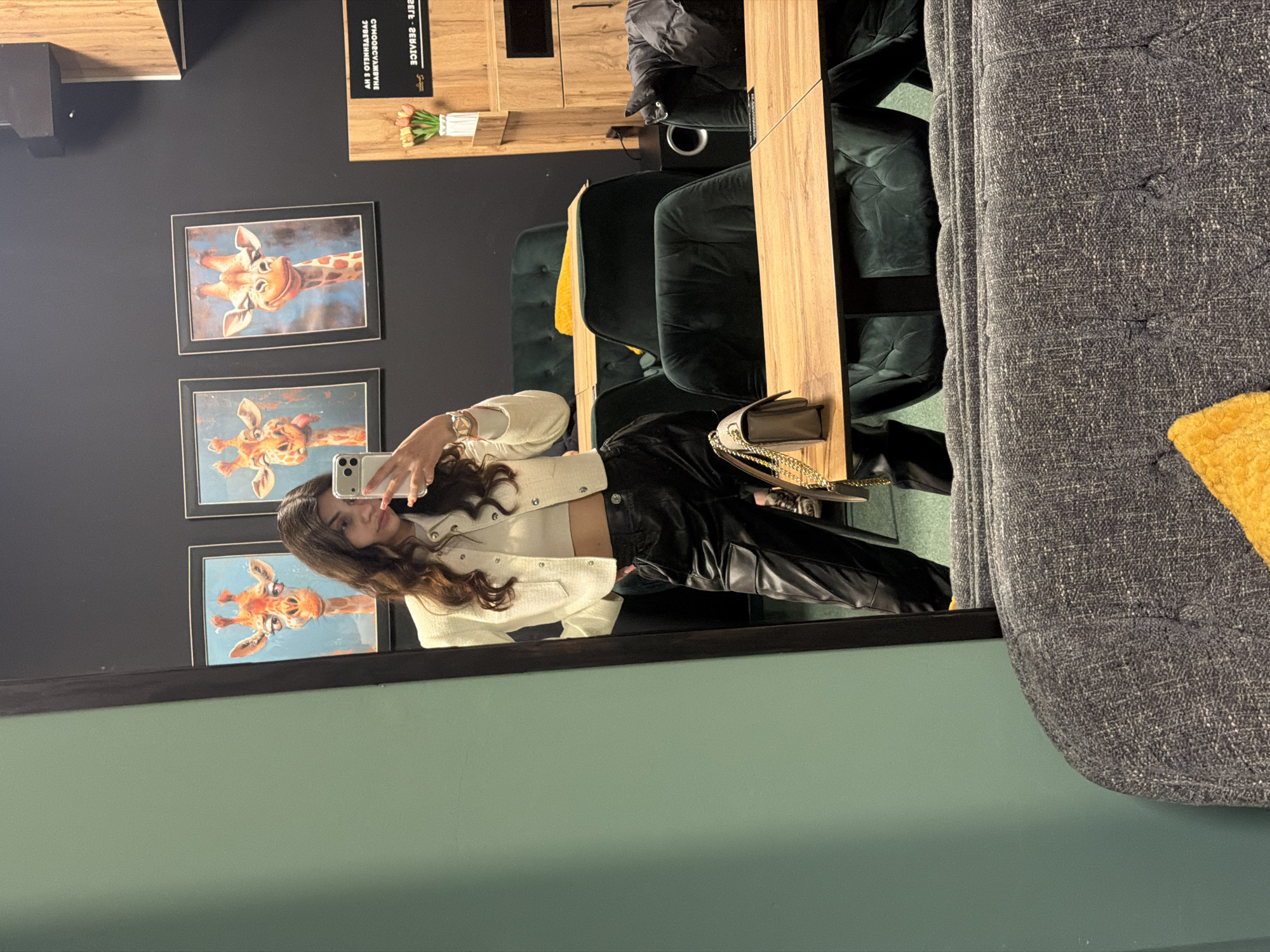 The image is a mirror reflection of a woman taking a selfie in what appears to be a cafe or lounge area. Three giraffe paintings are on the wall behind her. The woman is wearing a white jacket and black pants. In the background, there are dark green chairs and a wooden table. A grey upholstered sofa is visible on the right side of the mirror. The reflection of the wall is a light green color. The location might be in Veliko Tarnovo, Bulgaria, but there is no contextual information to verify this city.