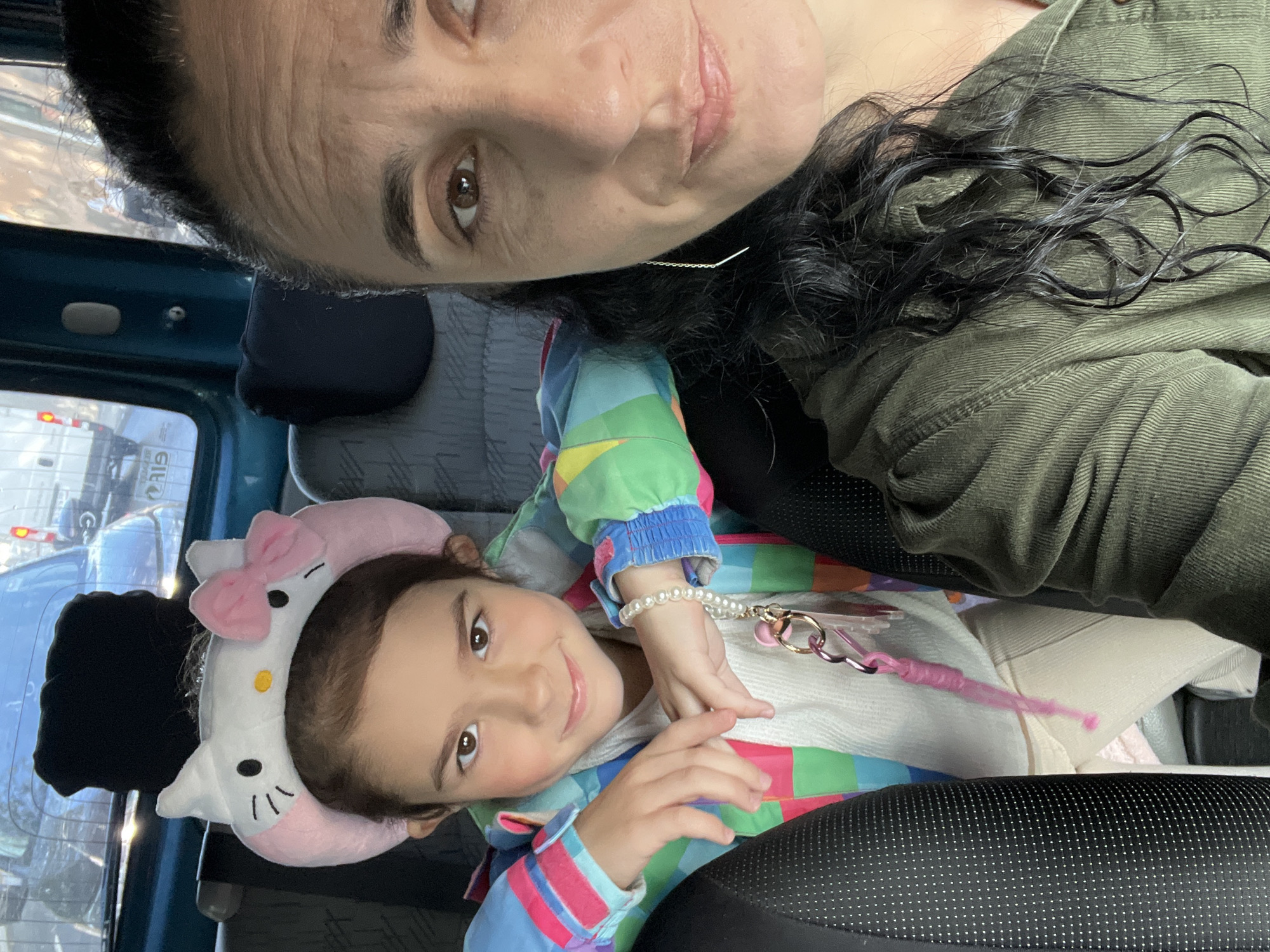 The image shows a selfie taken from inside a vehicle, with two people visible. On the left, a young girl is wearing a white and pink Hello Kitty headband and a colorful jacket. She has her hands clasped in front of her and is wearing a pearl bracelet with a pink keychain. To the right, a woman is in the foreground, her face partially visible. Both are seated in the back of what appears to be a van or bus, with dark seats and windows showing an outdoor environment. The setting is implied to be the city of Uzhhorod, Ukraine, based on contextual information. The lighting suggests it is daytime.