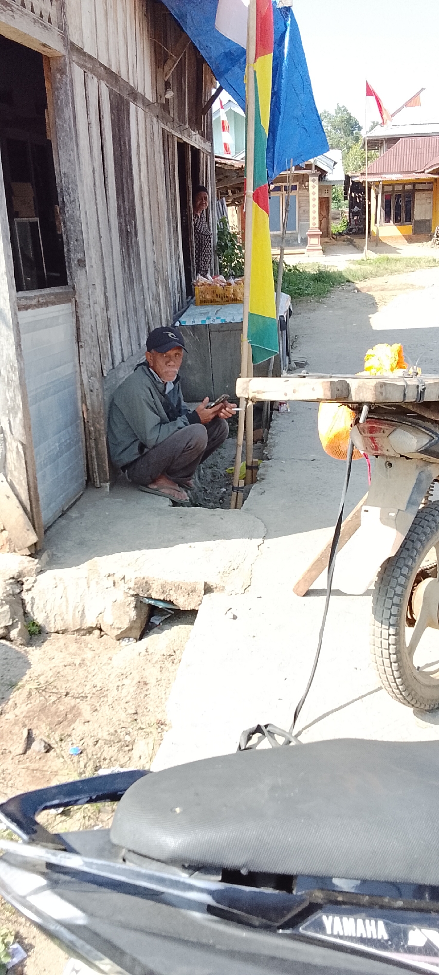 An elderly man wearing a dark baseball cap, a grey jacket, and brown pants is squatting on a concrete sidewalk. He is holding a mobile phone in his right hand and appears to be interacting with it. A black cable is draped over his left shoulder. The man is positioned in front of a wooden building with vertical planks. To his right, a part of a motorcycle is visible, including its rear wheel and seat with a "YAMAHA" logo. The background shows a dirt road and more buildings, some with red tile roofs, suggesting a rural or semi-urban environment. Flags, including one with blue, yellow, and green sections, are also visible. The scene is lit by bright sunlight, casting shadows on the ground. The area appears to be in Sukarame, Indonesia, given the contextual information.