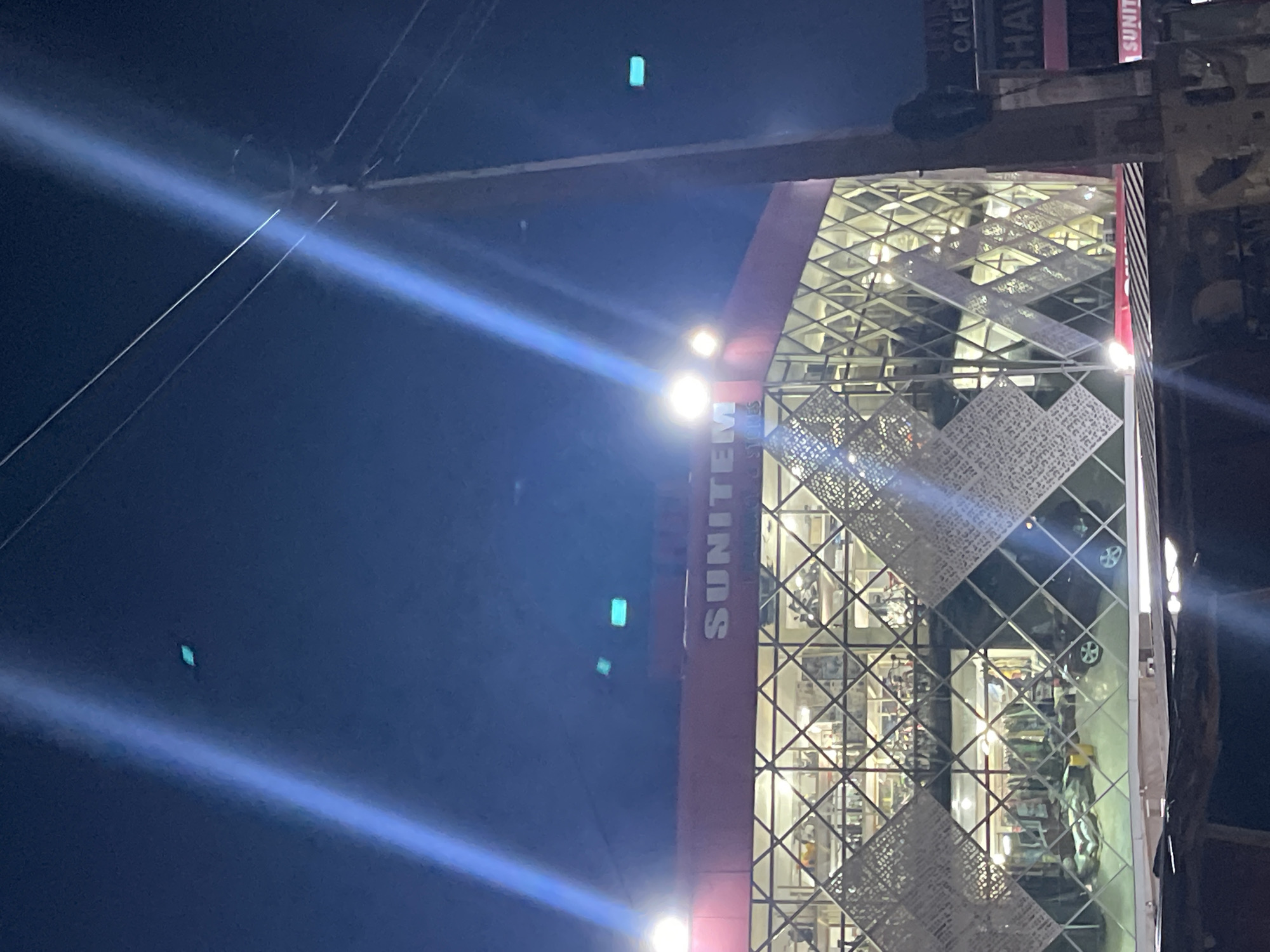 An upward-tilted nighttime view shows the exterior of a building with "SUNITEM STORES" in large, vertical block letters on a red surface. The building has a glass facade with a grid-like pattern overlaid with decorative diamond-shaped panels. The interior displays what appear to be shelves stocked with merchandise and parked cars. A bright light source originates from the building's facade, creating light rays extending into the dark blue sky. Power lines are visible diagonally crossing the image. Green artifacts are present in the sky, likely a digital imaging error. Signs including "CAFE SHAW" are present on the top right. The location is in Gwarinpa, Nigeria.