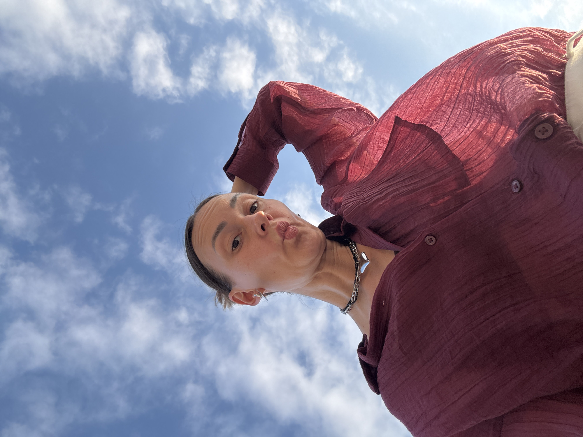 This media file is a low-angle, wide shot of a woman posing against a bright blue sky with scattered white clouds. She is wearing a loose-fitting, crinkled, deep red button-down shirt and a chunky silver chain necklace with a heart-shaped pendant. Her hair is pulled back, and she is puckering her lips in a playful gesture towards the camera. The lighting suggests it is daytime, likely sunny. The sky takes up a significant portion of the frame, indicating an outdoor setting. The image has a casual, selfie-like feel. The location is identified as Sochi, Russia.