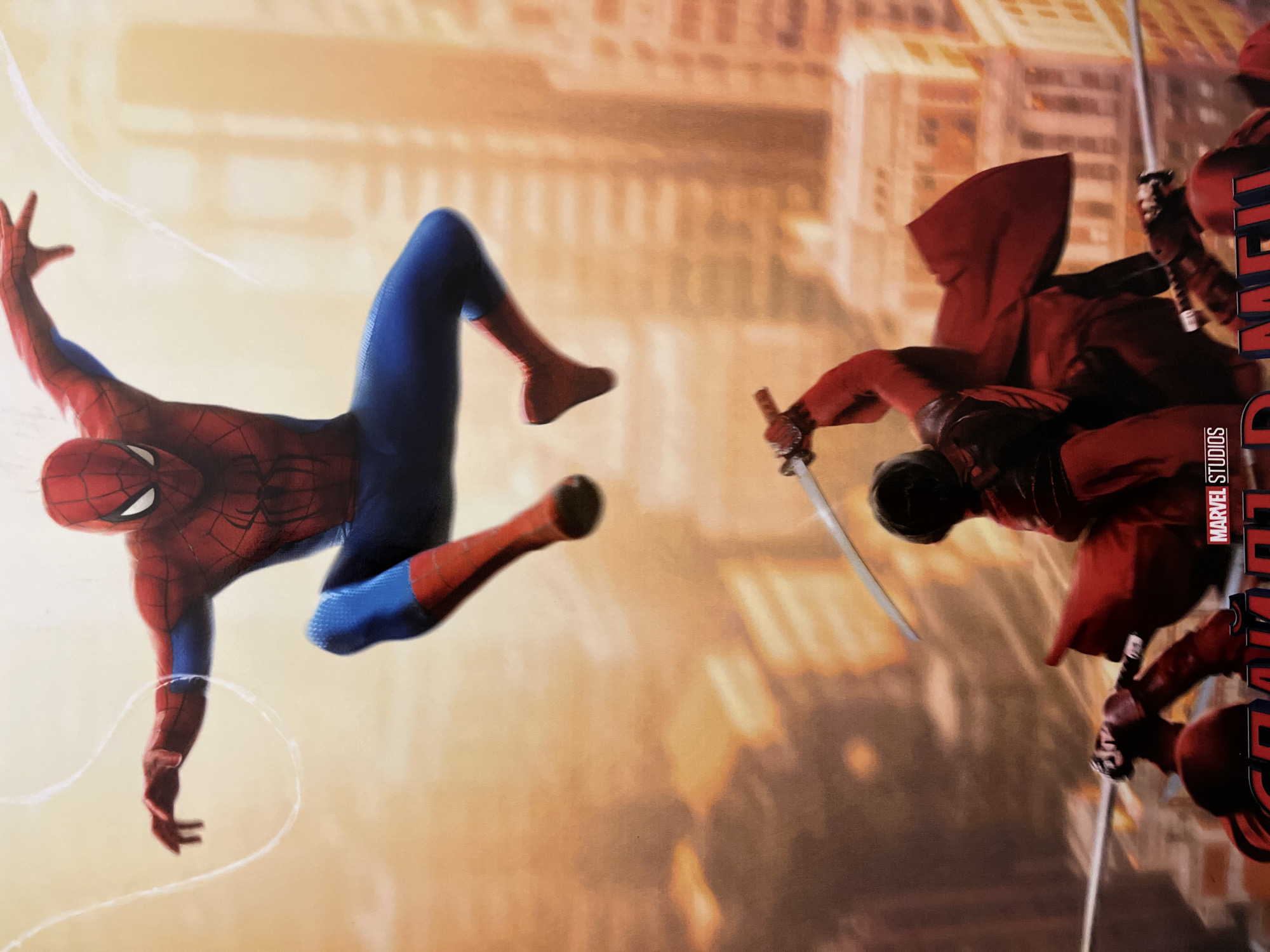 In an urban environment with blurred buildings in the background, Spider-Man, in his iconic red and blue suit, leaps through the air, a white web line trailing from his hand. Below him, a figure clad in red, with a flowing cape, wields a katana, poised to strike. Other figures in red can be seen with swords at the bottom of the frame. The Marvel Studios logo is visible on the poster.