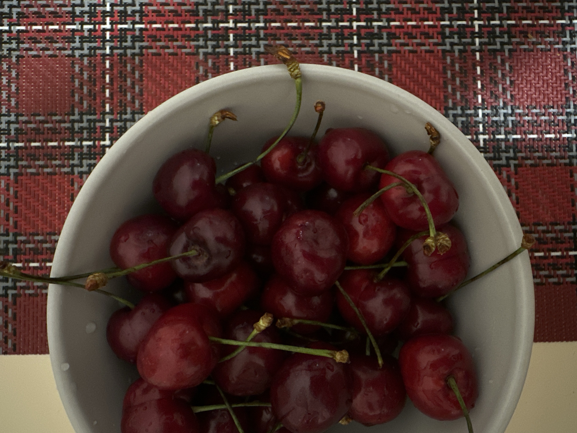 The image displays an overhead shot of a light gray, round bowl filled with numerous dark red cherries. The cherries appear fresh and plump, with a slight sheen and visible small water droplets on their surfaces, suggesting they may have been recently washed. Most cherries are still attached to their slender, green stems, some of which exhibit a slight brownish hue at their tips or where they connect to the fruit.
The bowl rests on a surface covered by a patterned placemat. The placemat features a woven, plaid-like design with intersecting lines and blocks in various shades of red, black, gray, and white. A plain, light beige or cream-colored surface is visible along the lower right edge, possibly indicating a tabletop.
No activity or event is taking place in the static composition. There are no visible people or specific location cues that would indicate the time of day, weather, or an exact geographical setting like Votkinsk, Russia. No visible text is present in the media file.