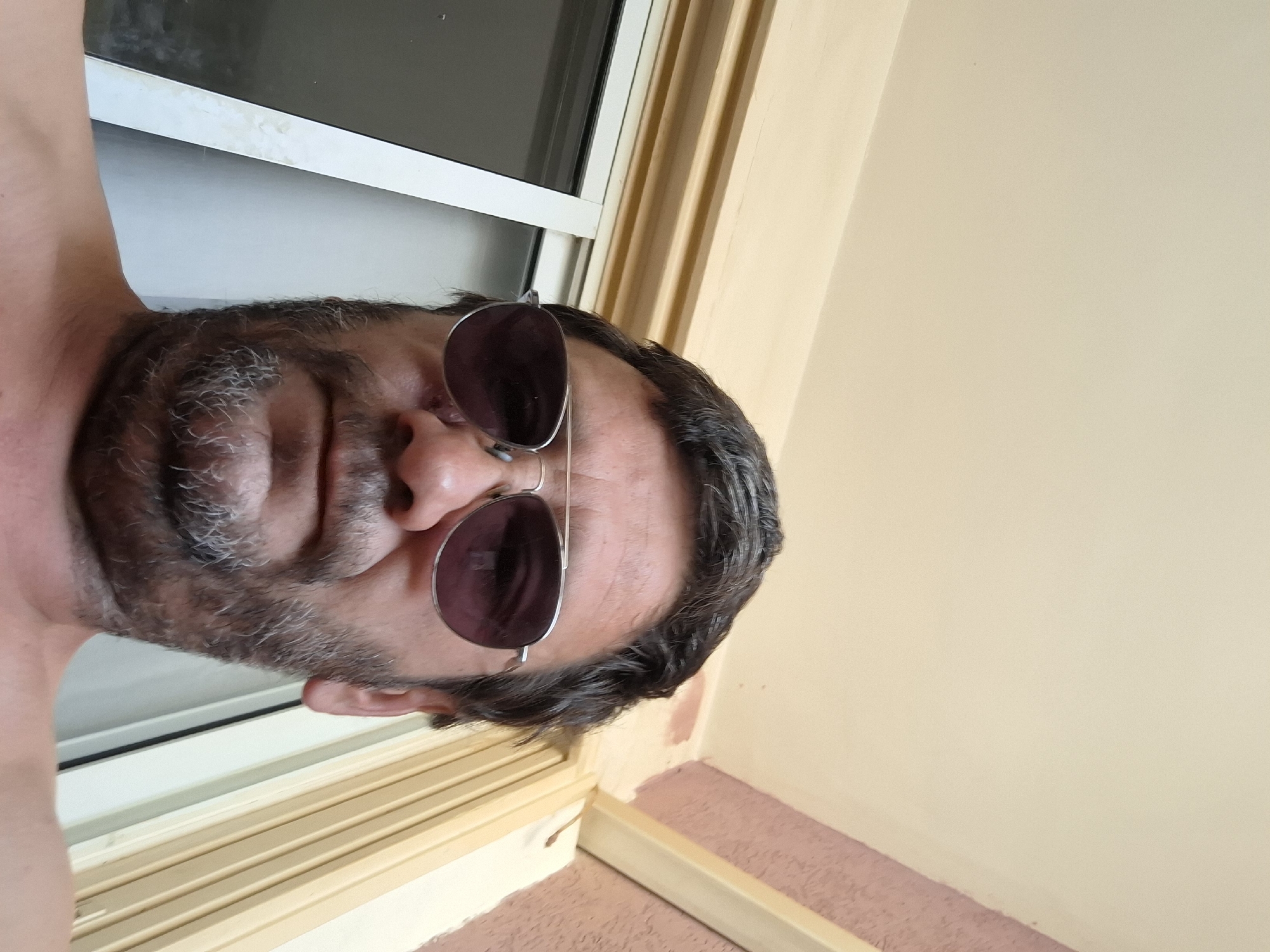 A man with dark hair and a beard wearing aviator sunglasses is shown from the chest up in an outdoor setting. The man is facing the camera, with his head tilted down slightly. Behind him, there is a beige ceiling with a pink textured wall and a window with vertical blinds. The image was taken in Yerocham, Israel.