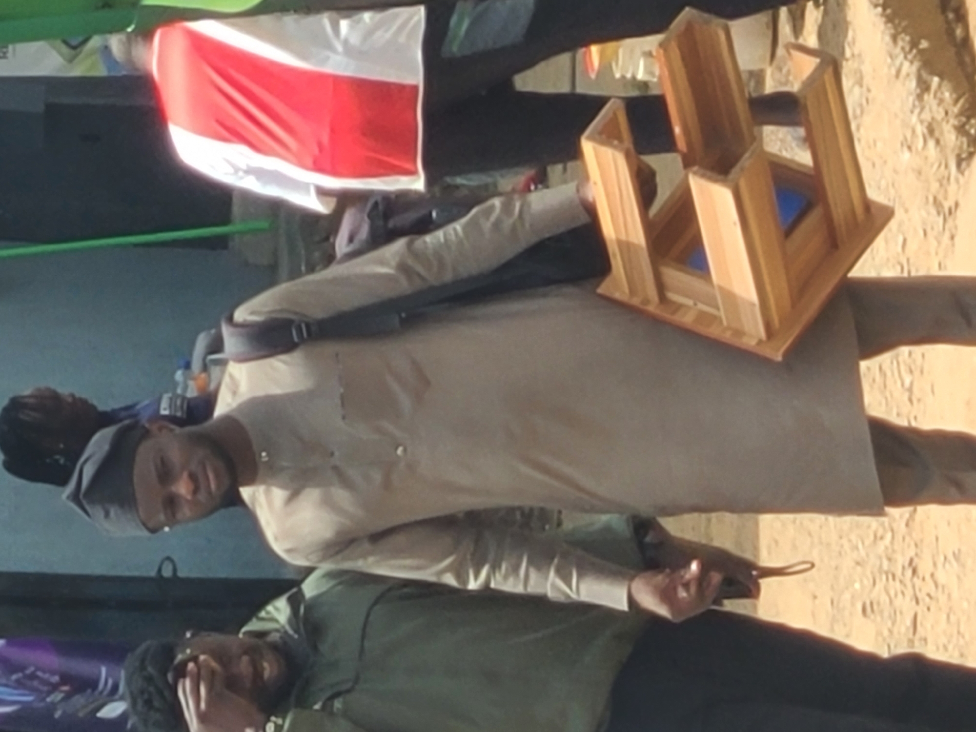 In Jos, Nigeria, a man wearing a tan traditional outfit and a dark kufi hat is seen holding a small, unfinished wooden stool. He is standing on a dirt road, with other individuals and partially visible structures in the background. To his left, another man in a green jacket is visible, with his hand on his forehead. In the background, to the right, a person in a red and white garment is also present. The environment appears to be an outdoor, possibly commercial or residential area with a rough ground surface.