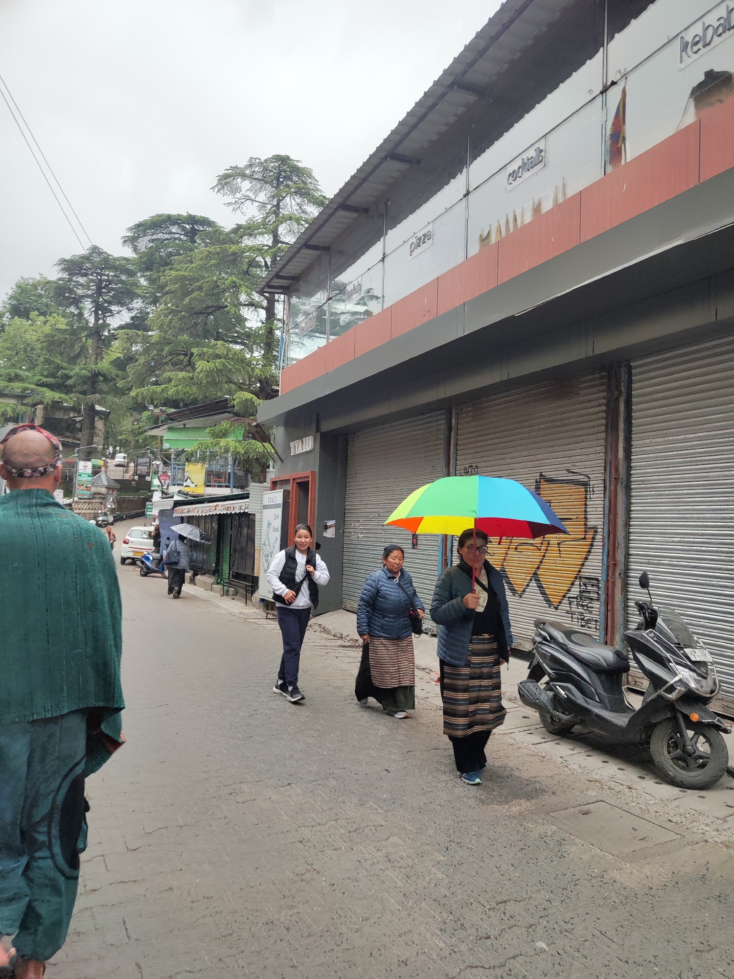 This media file captures a daytime street scene in Dharamshala, India, under an overcast sky. The paved street shows several pedestrians and parked vehicles.
In the foreground, a man with a receding hairline and a patterned headband stands with his back to the viewer, wearing a green garment. Moving along the street, a young woman in a white top and black vest smiles towards the camera. Further ahead, two women are walking; one holds a brightly colored rainbow umbrella, while the other walks beside her. Both women wear traditional-looking layered skirts. A black Suzuki Burgman scooter is parked on the right side of the street, next to closed shop shutters covered in graffiti, including a large yellow arrow and some indistinct writing.
The buildings lining the street feature modern facades on the upper levels, reflecting the overcast sky and surrounding trees. Visible signage on one building indicates "pizza," "cocktails," and "kebab," as well as "TYAULU Live Band." Lush green coniferous trees, characteristic of Himalayan regions, are prominent in the background and on the hillsides, suggesting a verdant mountain setting. The presence of the umbrella implies either recent rain or the expectation of it. The overall scene depicts typical daily activity in a bustling mountain town.