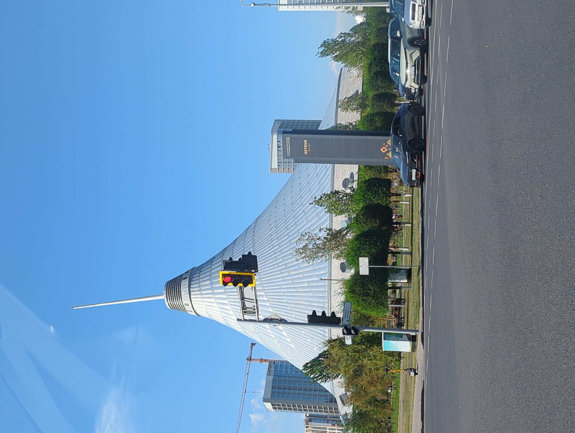 The image captures a street-level view in Astana, Kazakhstan, with the prominent Khan Shatyr Entertainment Center as the central architectural element. The structure is a massive, conical tent-like building with a metallic, silver exterior, topped by a tall, slender spire. A traffic light is visible in the foreground, displaying a red signal. Several cars are stopped at the intersection, including a dark-colored sedan, a white SUV, and another silver vehicle. Behind the Khan Shatyr, other modern buildings and construction cranes can be seen, suggesting ongoing urban development. The scene is set against a clear blue sky, with trees and manicured bushes lining the street, indicating a well-maintained urban landscape. A lone electric scooter is parked on the sidewalk, and bicycles are visible in the background, hinting at various modes of transportation in the city. The overall impression is one of a bright, sunny day in a modern, developing city.