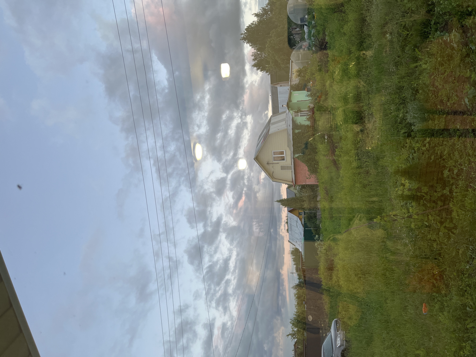 The image depicts a suburban scene in the village of Klyobushnevo, Russia, viewed through a reflective surface, likely a window. The foreground is dominated by tall, green grass and vegetation, with some orange debris visible in the lower left. A silver car is parked on the left side of the frame. In the mid-ground, several houses with pitched roofs are visible, along with fences and trees. One house is painted yellow with white trim, another is green with a silver roof, and a pink structure is partially visible on the far right. Several greenhouse structures are also present. The sky above is overcast with a mix of grey and white clouds, with hints of pink and pale blue suggesting either dawn or dusk. Three circular, illuminated fixtures are reflected in the viewing surface, appearing to be ceiling lights. Power lines are visible stretching across the upper portion of the image. The overall impression is one of a rural or semi-rural residential area during a transitional time of day.