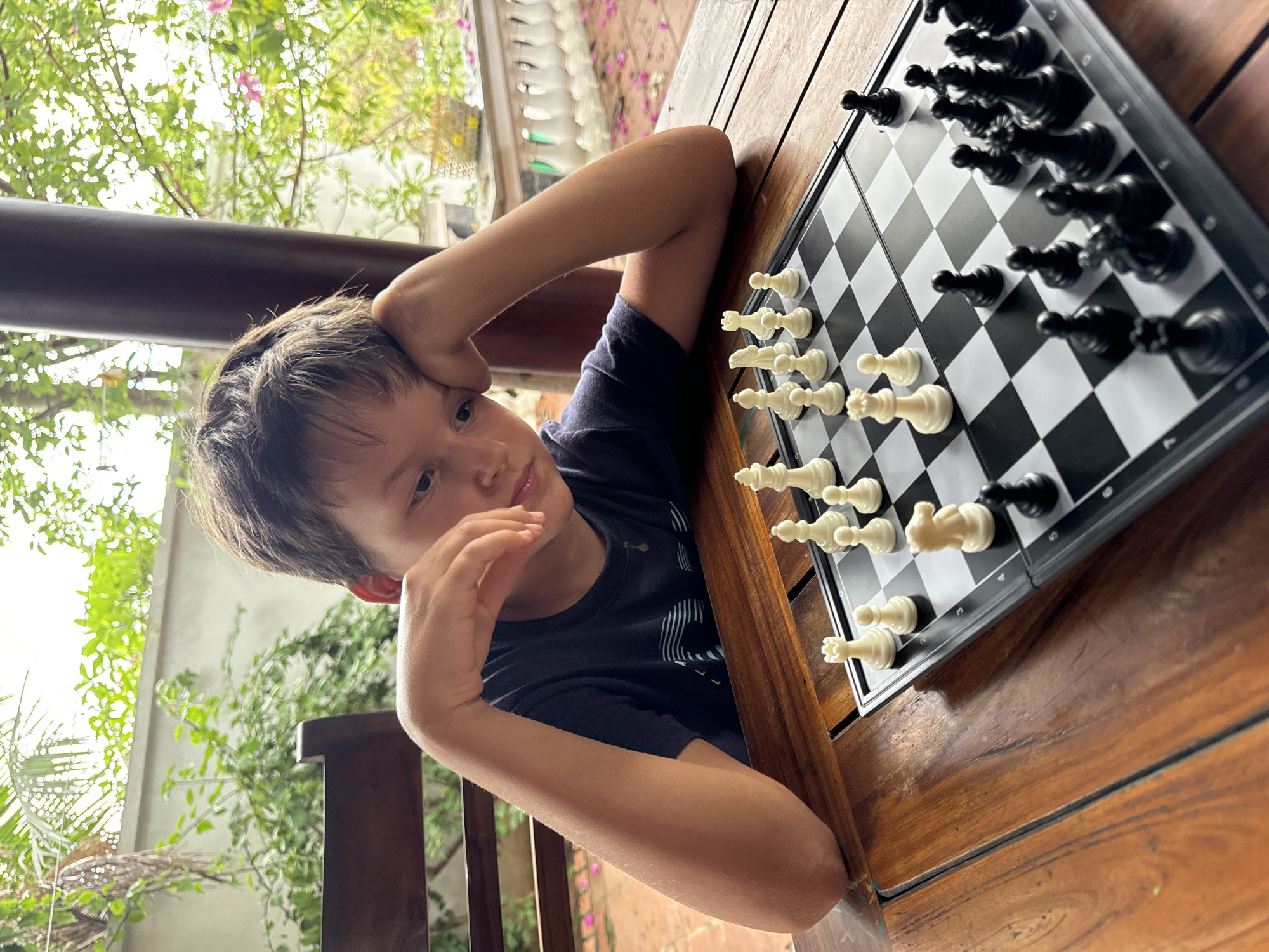 A young boy sits at a wooden table with a chessboard on it, resting his head on his hands as he looks at the board. The chessboard is set up with white pieces on the near side and black pieces on the far side. The table appears to be located outdoors, possibly on a balcony or patio, with foliage and a railing visible in the background. The location is likely Mũi Né, Vietnam.