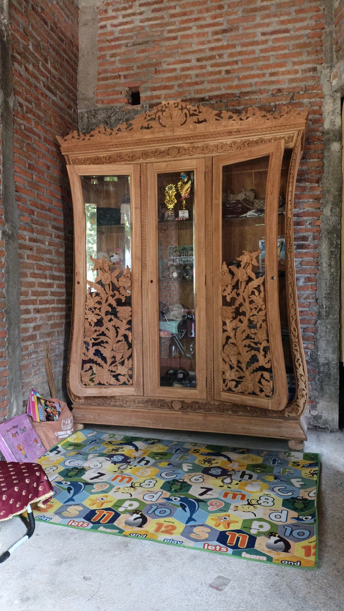 The image shows a large, ornately carved wooden cabinet with glass doors, positioned against a brick wall. The cabinet is filled with various items, including trophies, what appear to be stuffed animals, and other decorative objects. In front of the cabinet is a colorful play mat with letters, numbers, and cartoon animals. To the left of the cabinet, there are some books and a small red stool with a patterned cloth. The environment appears to be an indoor or semi-outdoor space, with a concrete floor and exposed brick walls. The overall scene suggests a home or a shop in Cikal, Indonesia.