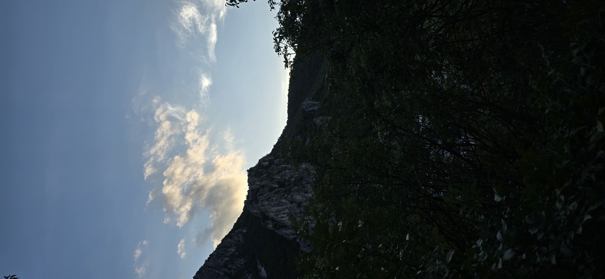 The image depicts a dramatic mountain landscape with a clear blue sky and wispy clouds. The sun is low in the sky, casting a warm glow on the clouds and illuminating the side of the mountain. The mountain itself is steep and rocky, with dark green foliage covering its lower slopes and a thick canopy of trees in the foreground. The scene is captured in a vertical orientation, creating a sense of depth and scale. The time of day appears to be late afternoon or early evening, given the soft lighting and the angle of the sun. The weather seems to be fair and clear, with the exception of a few scattered clouds. The location is implied to be Vratsa, Bulgaria, a region known for its mountainous terrain and natural beauty. The overall mood of the image is one of serenity and awe, with the grandeur of nature taking center stage. There are no people or specific objects visible in the image, allowing the viewer to focus solely on the natural elements.
