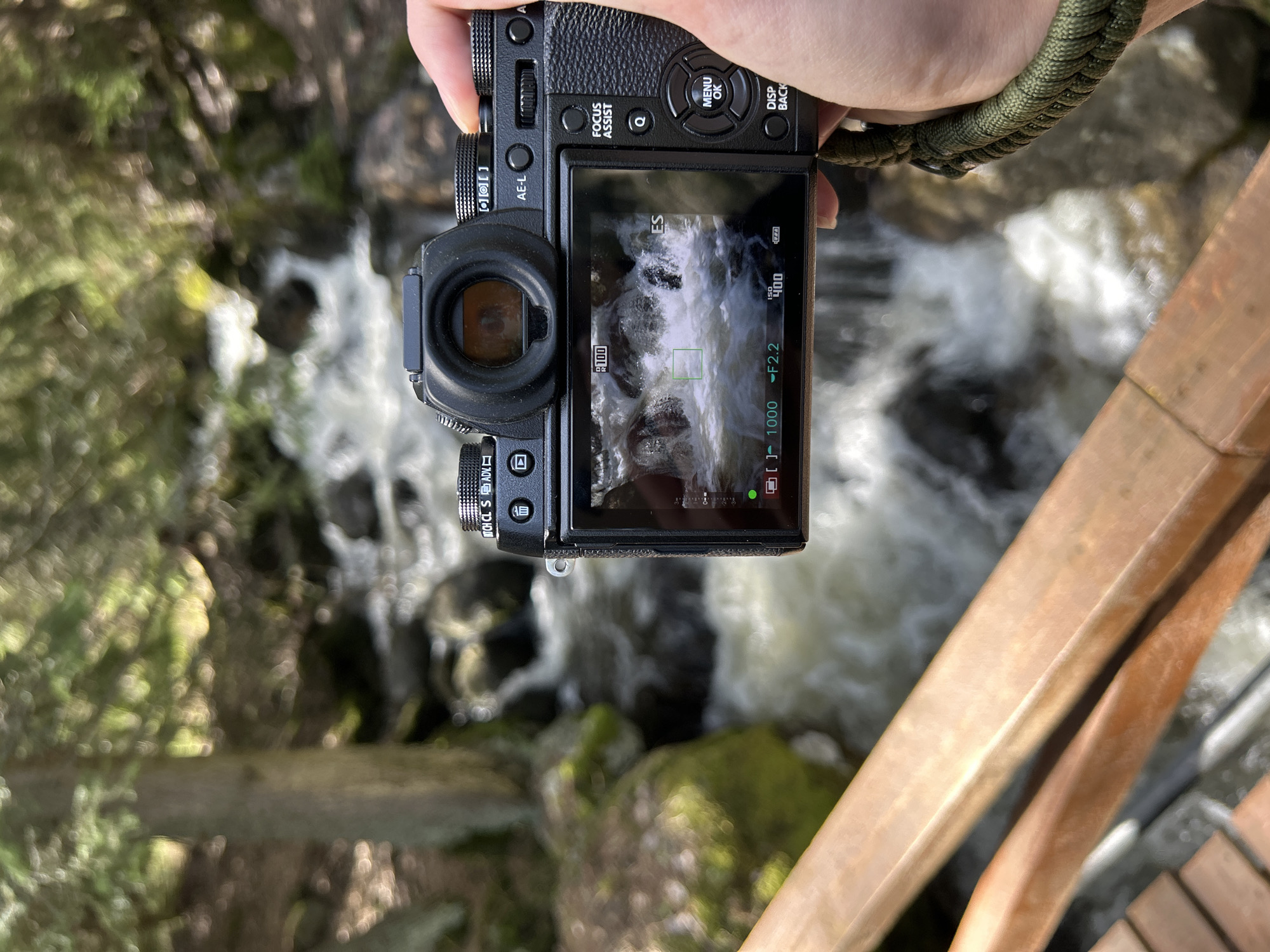 A photograph shows a camera being held by a person, pointed towards a waterfall. The camera's screen reflects the scene it's capturing - the waterfall and surrounding rocks. The hand holding the camera wears a green paracord bracelet. A wooden railing is visible in the lower right of the shot. The location appears to be a natural area, possibly a park or wilderness setting, with trees and moss-covered rocks visible, although the exact city and state are not identifiable from the image. The interaction is the act of taking a photograph.