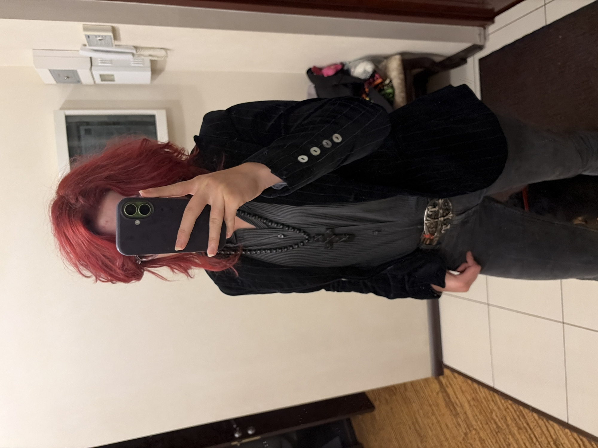 A person with bright red, voluminous hair is standing indoors and taking a mirror selfie with a smartphone. They are wearing a dark, pinstriped blazer over a grey, vertical-striped shirt. A beaded necklace with a large cross pendant is visible. The person also has on grey jeans and a decorative belt with a silver buckle. In the background, a white intercom system is mounted on the wall, and to the left, a doorway and a wooden floor are visible. The floor is tiled with light-colored squares.