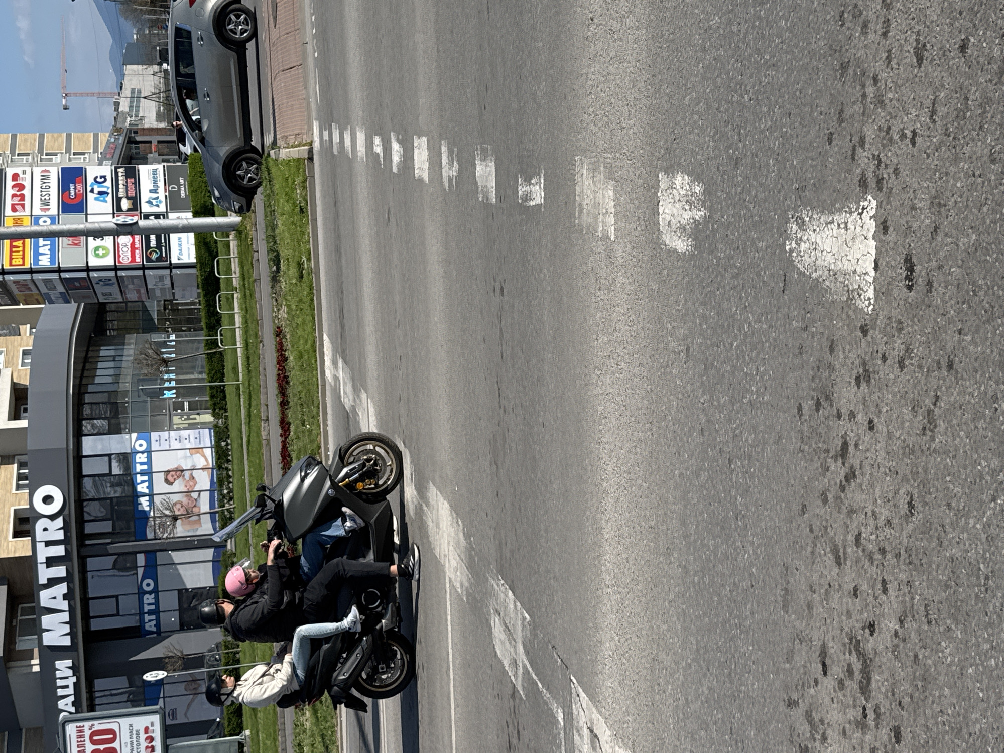 The image shows a street scene in Plovdiv, Bulgaria. A scooter with two riders, one wearing a pink helmet and the other wearing a white jacket, is stopped on the side of the road near a building with the name "MATTRO" displayed prominently. A car is parked along the sidewalk in the background. The street has a faded arrow marking and dashed white lines indicating lanes. Billboards with various company logos and advertisements are visible in the background, including logos for "BILLA", "MAT", "WESTGYM", "ATG", and "Armeec".