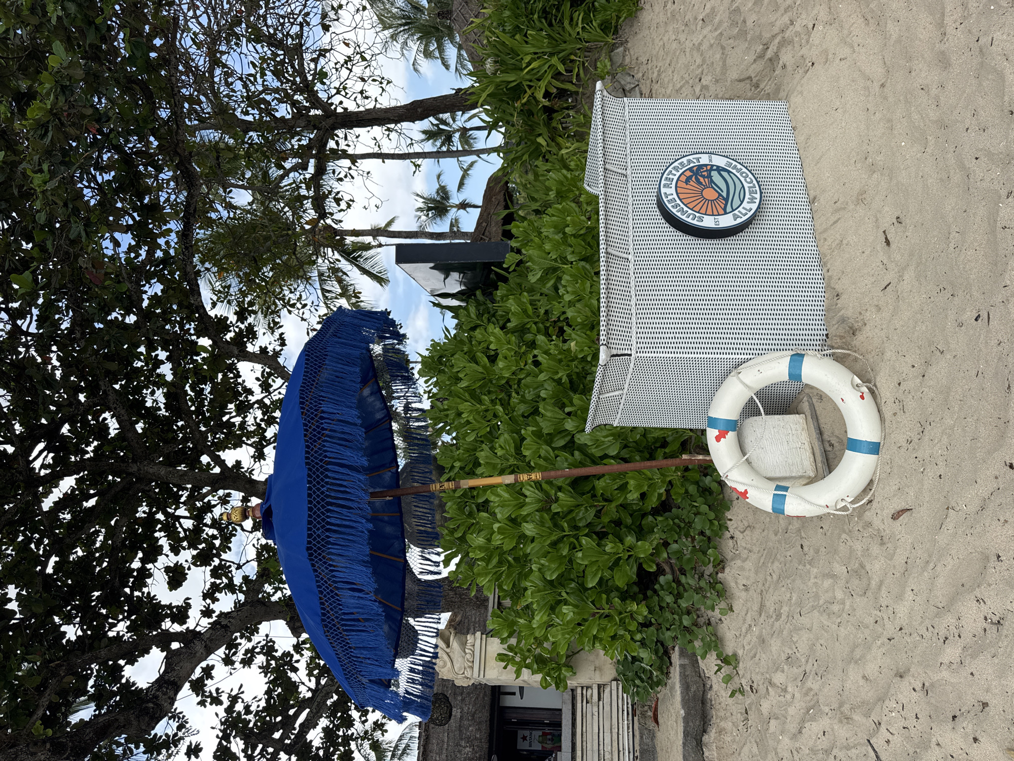 The image depicts a beach scene in Kuta Selatan, Indonesia. A white, metal chair with a circular "Sunset Retreat" logo on its back stands on the sand. A white life preserver with blue stripes hangs on the side of the chair. To the left of the chair, partially obscured by green foliage, is a blue fringed umbrella attached to a wooden pole. The scene is shaded by a large tree to the left, while palm trees are visible in the background. The setting appears to be a resort or beach club.
