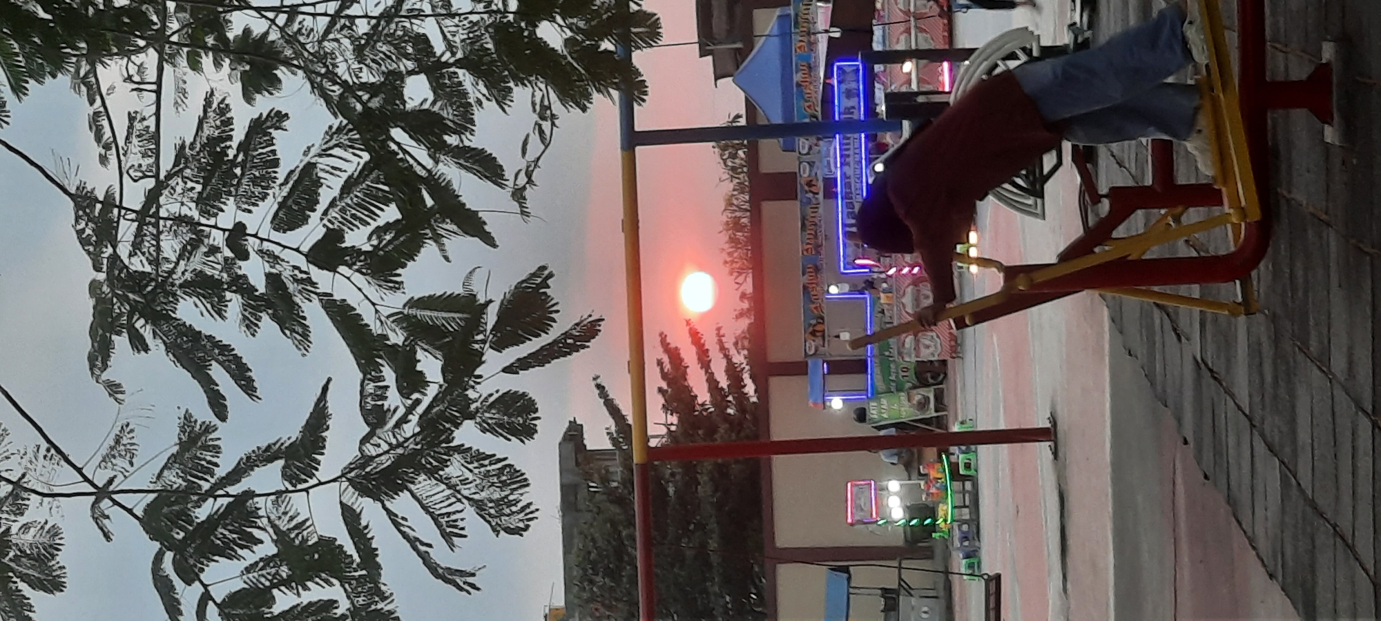 This is a medium-shot, vertical-orientation, eye-level photograph taken at sunset in a public park or recreational area in Kanigoro, Indonesia. The foreground features a red and yellow exercise machine, with a person wearing a maroon long-sleeved shirt and blue jeans using it, positioned facing away from the camera. Their arms are extended, and they appear to be engaged in a leg-pressing or similar movement. In the midground, there are several food stalls or kiosks with colorful lights and signage, suggesting a lively evening atmosphere. Behind the stalls, there is a building with a concrete wall and a section of what appears to be a roof or upper structure. The background is dominated by the sky, which is a pale gray with a bright, hazy orange sun setting near the horizon, casting a warm glow. Overhanging the scene from the top are the branches and leaves of a tree, silhouetted against the sky. The ground is paved with interlocking dark gray tiles. The overall impression is of an active outdoor space during the twilight hours.