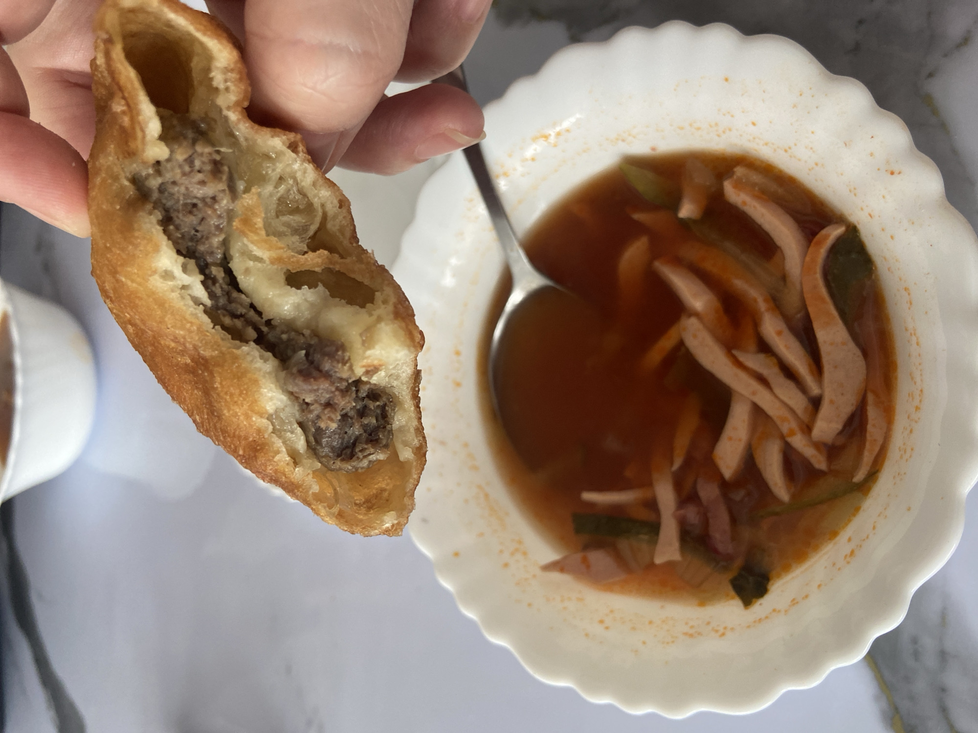 A hand is holding a half-eaten, fried pastry filled with ground meat and cheese over a white bowl of soup. The soup appears to be a broth with sliced sausage and some green vegetables. The background is a white, textured surface. There is no indication of the location being in Усмангали, Russia.