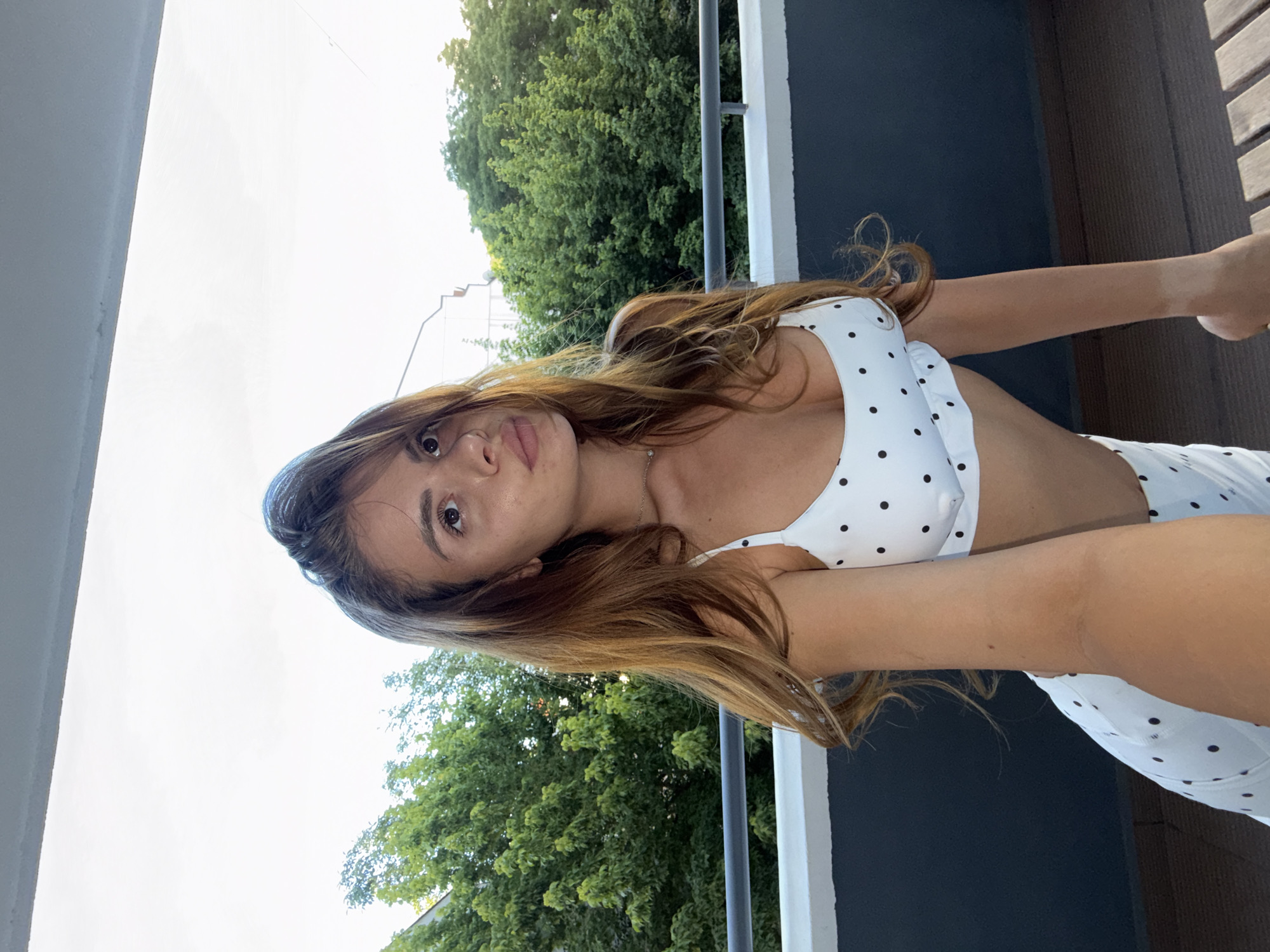 A young woman with long, wavy brown hair is standing on a balcony, holding up her phone to take a selfie. She is wearing a white bikini with black polka dots. In the background, lush green trees and a bright sky are visible. The balcony appears to overlook a residential area.