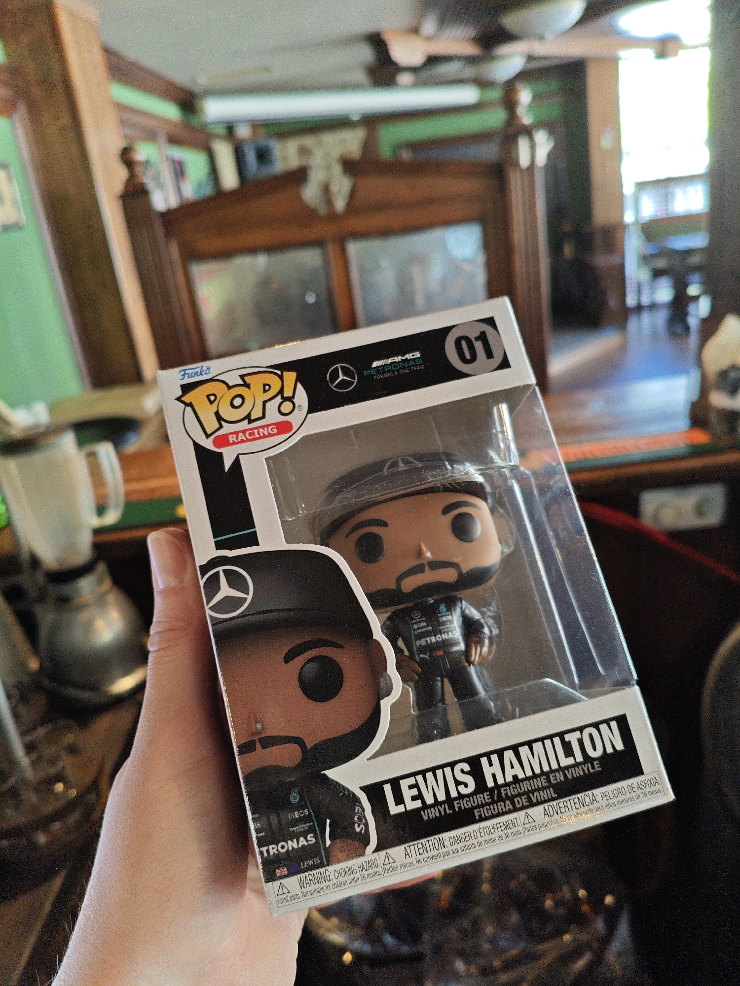 A hand holds a Funko Pop! vinyl figure of Lewis Hamilton, a Formula 1 driver. The figure is encased in its original clear plastic packaging, which has "Funko Pop! RACING" printed on the top. The figure depicts Hamilton in his racing suit with the Mercedes-AMG Petronas Formula One Team logos. The background is blurred and appears to be an indoor setting with wooden furniture and green walls, possibly a room in a house or a establishment. There is no visible indication that this image was taken in Kyiv, Ukraine.