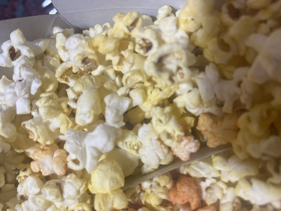 The image presents a close-up, high-angle view of a quantity of popcorn contained within a light-colored, likely cardboard, box visible along the right edge. The popcorn consists predominantly of white and pale yellow kernels, with several pieces exhibiting an orange-red seasoning, suggesting a flavor additive. A slender, light-colored object, consistent with a popcorn scoop or divider, is partially visible diagonally embedded in the popcorn, extending from the lower-left to the upper-right. No individuals or specific actions are depicted. The scene is consistent with an indoor commercial establishment, such as a cinema or entertainment venue in Mexico City, Mexico; however, no specific identifying landmarks or features of Mexico City are visible within the frame.