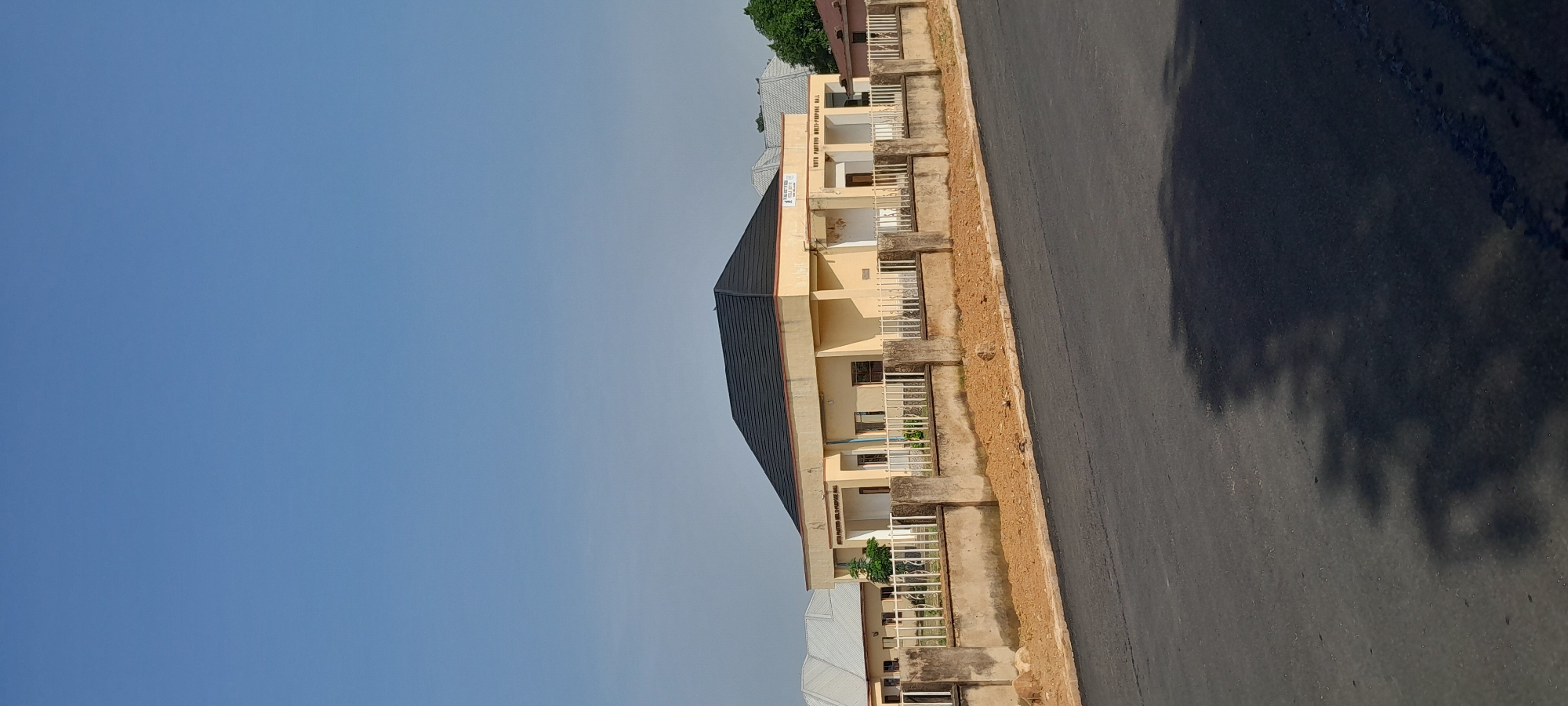 A three-story building with a dark, pitched roof stands behind a low concrete wall and a white metal fence. The building is painted a light yellow color and has several windows and a doorway. A sign above the entrance reads "YOUTH PASTORS MULTI-PURPOSE HALL". The building is located in Jalingo, Nigeria. The image is taken from a slightly elevated angle, showing a paved road in the foreground with shadows of trees cast on it. The sky is clear and blue.