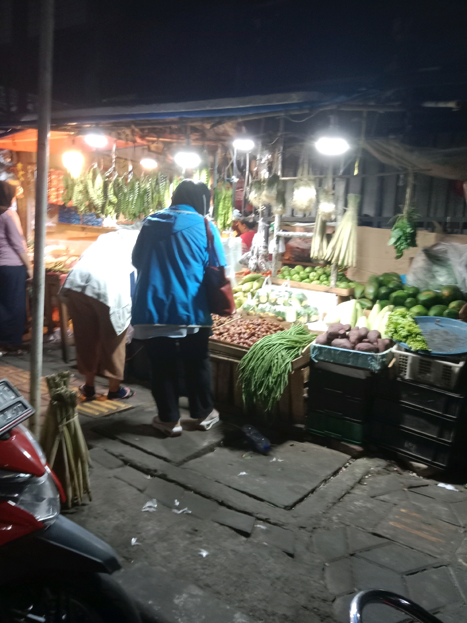 At night, a street-level market scene unfolds in the city of South Tangerang, Indonesia. The market is illuminated by hanging bulbs, casting a bright glow on the produce displayed. A woman in a blue jacket and dark pants, along with another person in a white shirt and brown pants, are seen browsing the goods. Stalls are laden with various vegetables and fruits, including long green beans, what appear to be potatoes, green leafy vegetables, and other produce. The ground is paved with textured tiles, and a red motorcycle is parked on the left side of the frame, partially obscuring a license plate. Bundles of dried grass or reeds are also visible near the motorcycle. The overall impression is that of a busy, open-air market at dusk or night.
