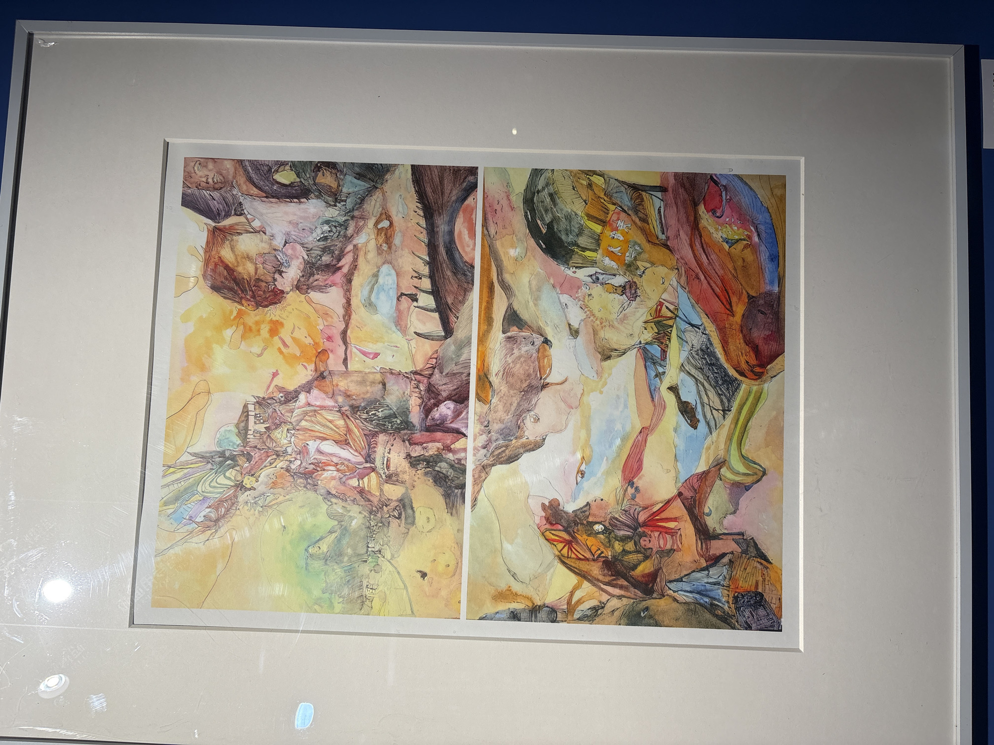 A framed abstract watercolor painting hangs on a wall, likely in a gallery or museum, within the Nan'an District of China. The painting is divided into two panels, both depicting surreal and organic forms in a palette of yellow, orange, red, blue, and green hues. The left panel contains more yellow tones and the right more reds. Each panel consists of a riot of lines, washes, and shapes, resembling both natural landscapes and anatomical features. The frame is a wide white molding. The wall behind the painting is a dark blue.