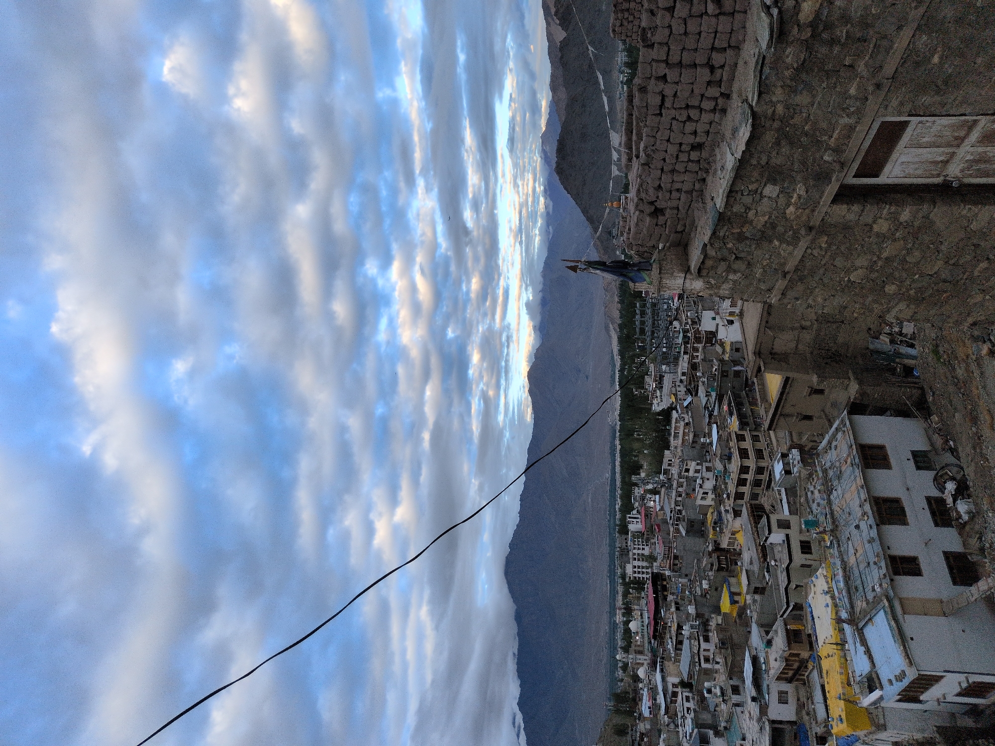 An elevated shot captures the town of Leh, India, under a dramatic, cloudy sky. In the foreground, a rough stone wall of an ancient structure dominates the right side, featuring a weathered wooden door. On top of this structure, a single figure stands, partially obscured, beside a pole with prayer flags. Beyond this, a dense collection of buildings with flat roofs, some covered in yellow tarpaulins, sprawls across the landscape. In the distance, a vast mountain range, shrouded in mist, forms a formidable backdrop to the settlement. A black wire bisects the upper portion of the image, stretching from the left edge. The overall atmosphere is muted, suggesting either early morning or late evening light.