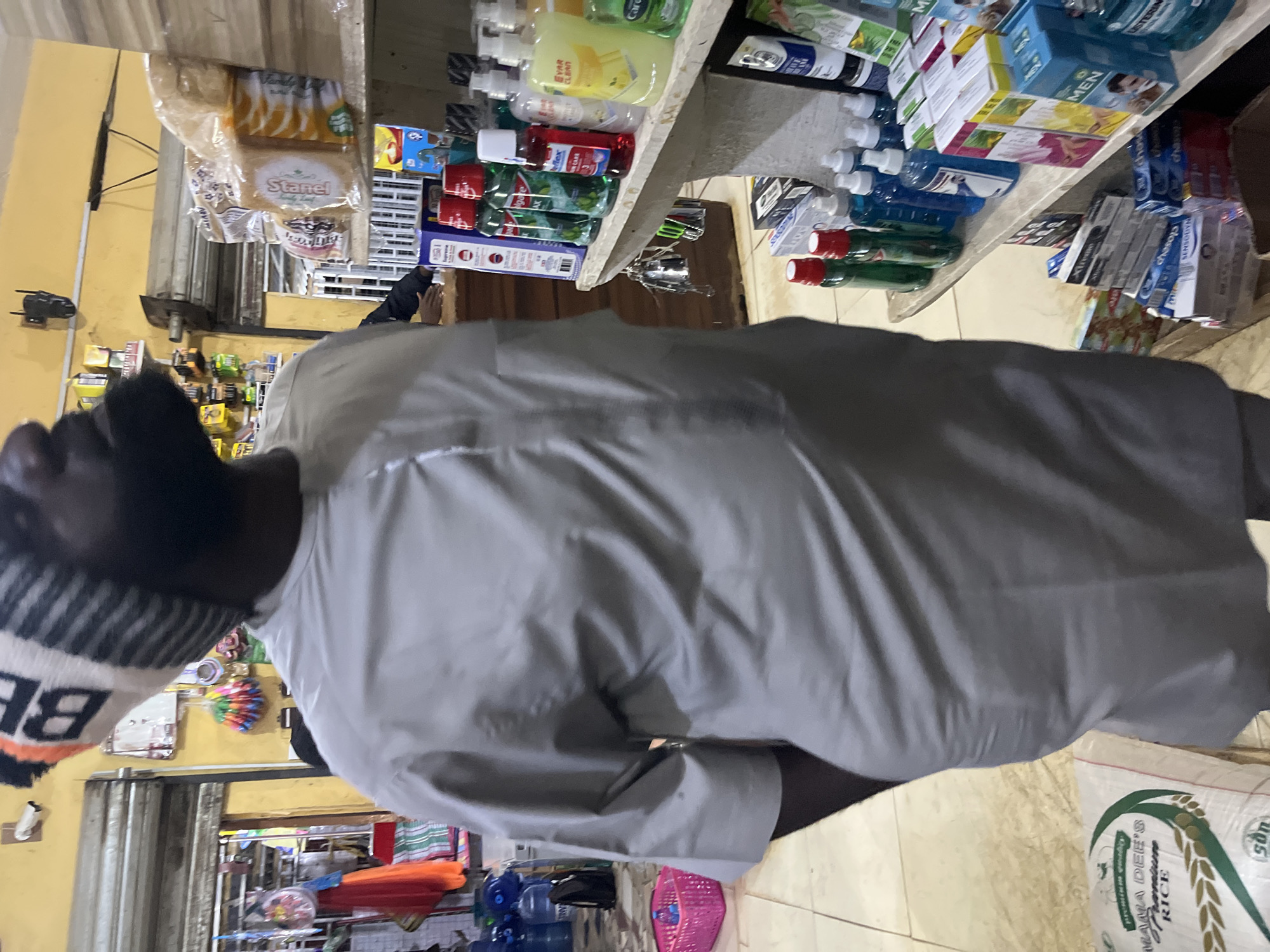 The image shows a man wearing a grey traditional outfit and a beanie standing in a shop. He is facing away from the camera and appears to be looking up. The shop is filled with various products, including bottled water, canned goods, hygiene products, and packaged food items. In the background, there is a closed shutter and more shelves stocked with goods. The man's posture and the general ambiance suggest he is a customer browsing or making a purchase. The location is identified as Bukuru, Nigeria.