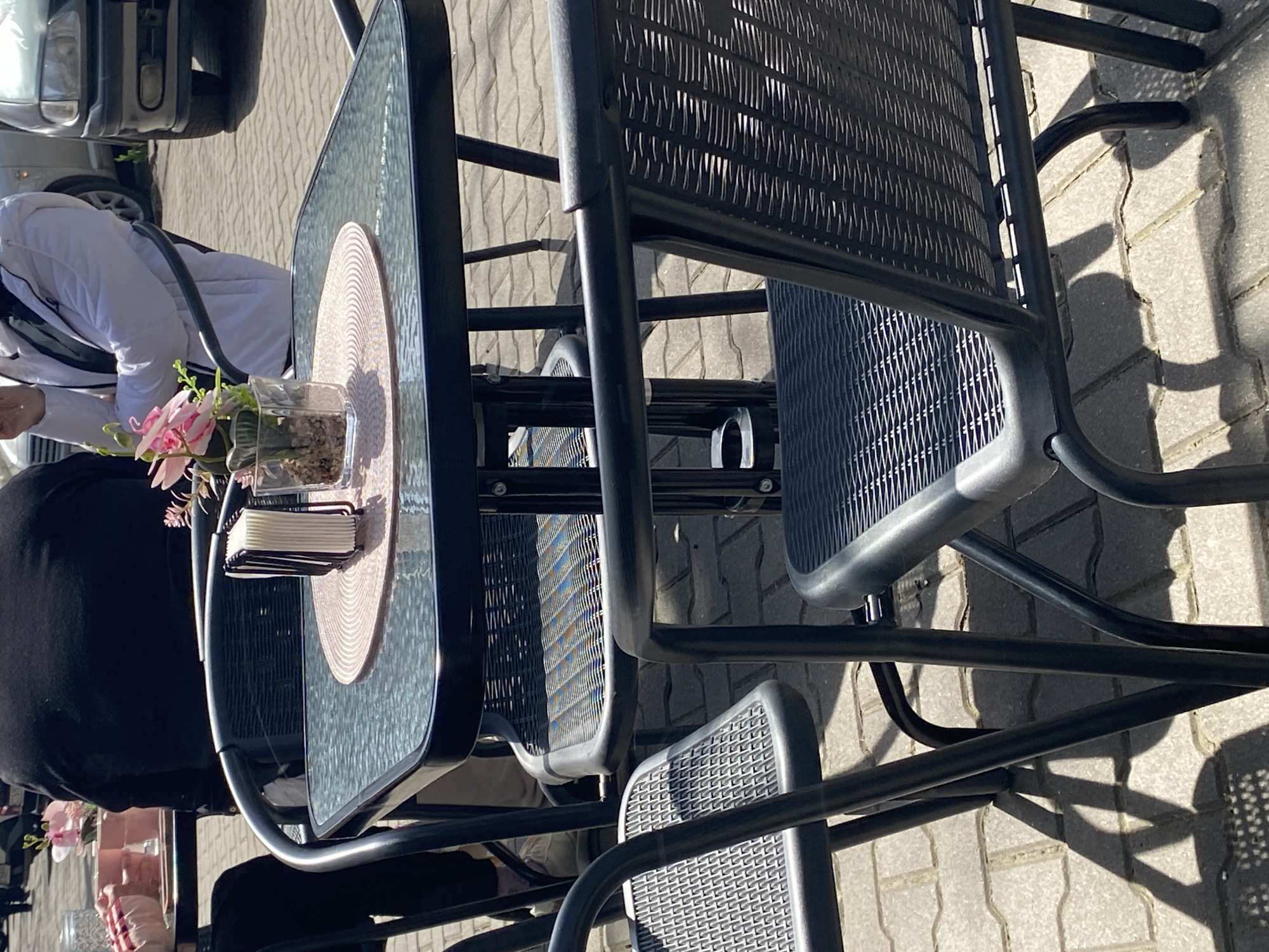 An outdoor café scene is depicted on a sunny day in Stara Zagora, Bulgaria. In the immediate foreground, a black metal-framed table with a clear glass top is centrally positioned. It features a round, pink woven placemat on which a black, vertical napkin holder containing white paper napkins and a small, clear glass vase holding pink artificial flowers and green foliage, set in decorative gravel, are arranged. Several black metal-framed chairs with dark gray, woven plastic seats and backrests surround this table; one chair is in the lower left foreground, and parts of others are visible to the right and behind the table.
To the left of the central table, a person with their back to the viewer wears a black top, with their left arm resting on an adjacent table where a pink cup is visible. To the right, another individual, partially obscured by the central table, wears a white padded jacket over a black garment, with their hands positioned near their face.
The ground is paved with light-colored rectangular pavers, casting distinct shadows from the chairs and table, indicating direct sunlight. In the background, beyond the seating area, several vehicles are present, including the front grille of a silver or white car and the front section of a dark gray/green car with its headlight and bumper visible.
