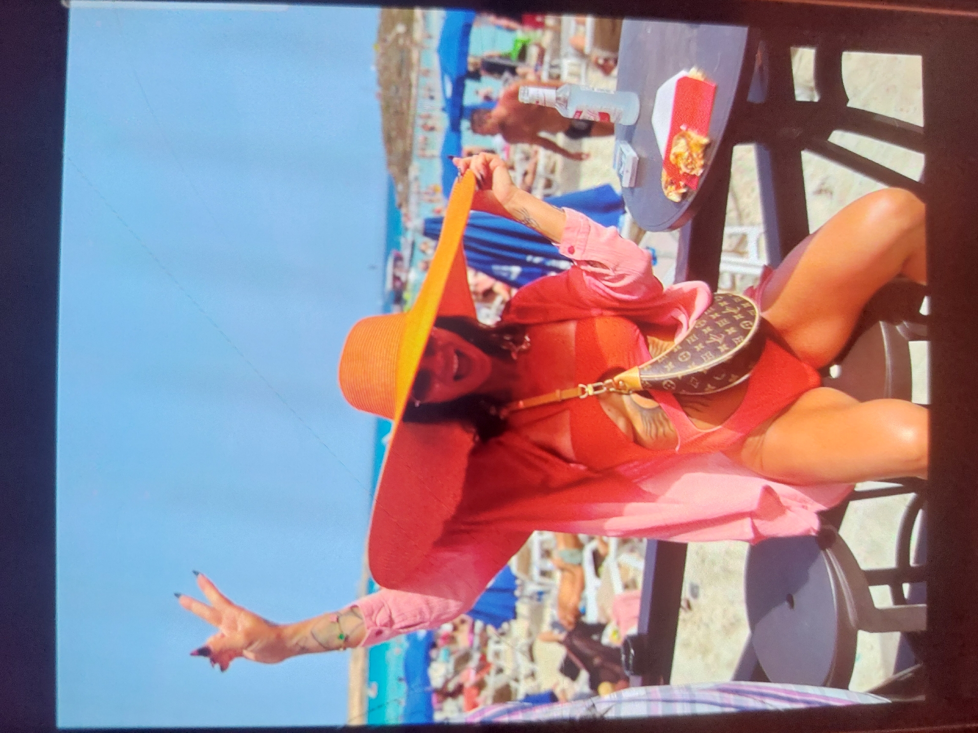The image captures a woman posing at a table on a sunny beach in Tsarevo, Bulgaria. The main subject is a woman with tanned skin, wearing a large orange wide-brimmed straw hat, a light pink long-sleeved shirt unbuttoned over a red/orange bikini top, and a distinctive Louis Vuitton monogrammed shoulder bag. She has dark nail polish and visible tattoos on her arms. Her right hand is holding the brim of her hat, while her left hand is raised in a gesture.
On the dark-colored table beside her, there is a white bottle, possibly of water or spirits, and a plate holding food that appears to be fries or similar items, served on a red and white checkered liner.
The background reveals a bustling beach scene under a clear blue sky. Numerous blue beach umbrellas and loungers are visible, occupied by many beachgoers. In the mid-ground, a shirtless man is walking on the sand. The overall setting suggests a vibrant, warm, and sunny day at a popular beach destination. The bright lighting indicates it is daytime, likely mid-morning or afternoon.