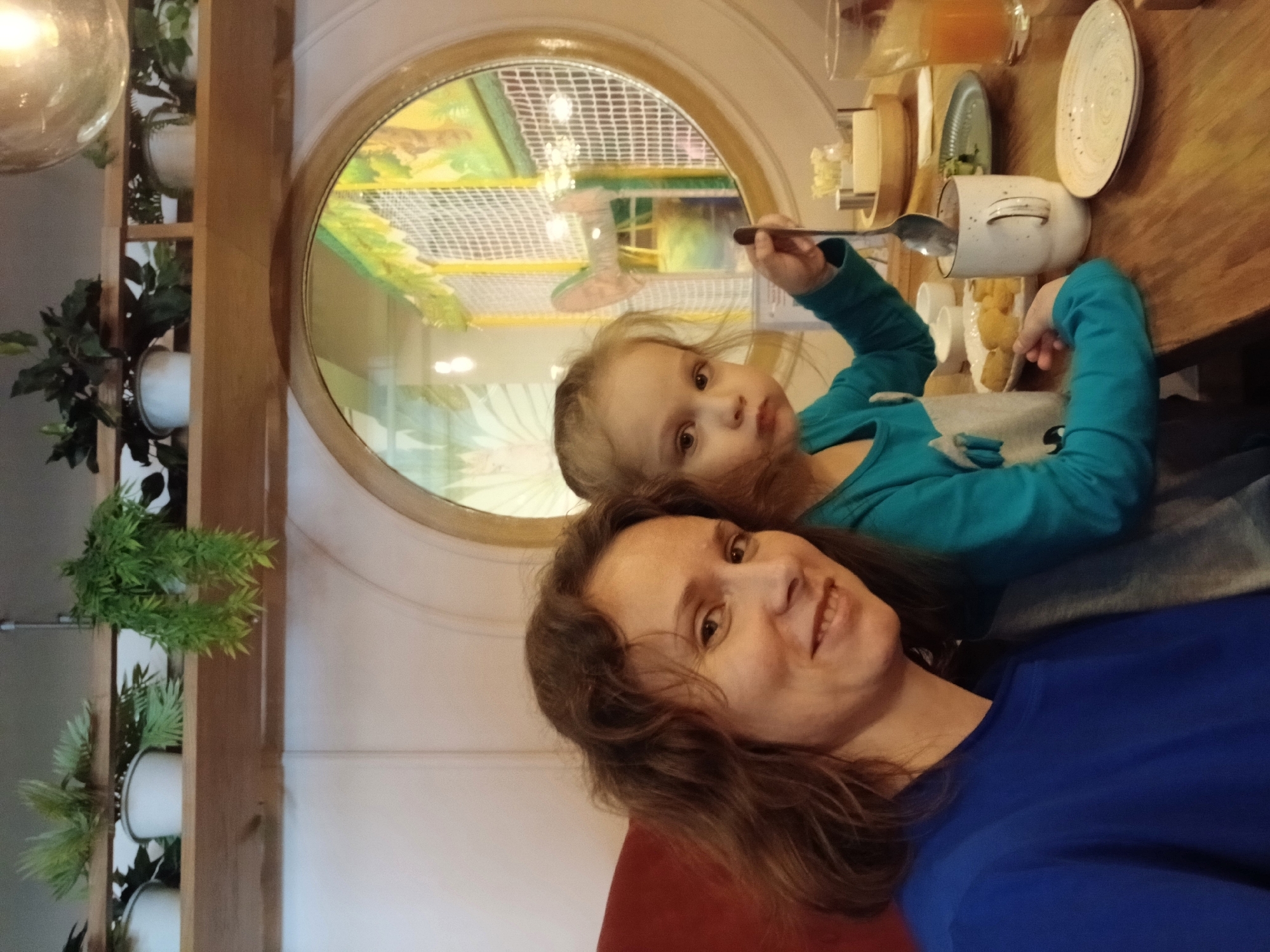 A woman and a young child are seated at a wooden table, indoors. The woman, on the left, has brown, wavy hair and is smiling. She is wearing a blue shirt. The child, on the right, has blonde hair and is wearing a teal shirt with a white graphic. The child is holding a spoon, which is inside a white mug. In front of them on the table are plates of food, a glass of orange juice, and other tableware. Behind them, a round window offers a view into a colorful play area, suggesting the location is a restaurant or cafe with a children's play zone. Plants are visible on shelves above and behind the subjects. The overall ambiance is casual and family-friendly. The image was taken in Yekaterinburg, Russia.