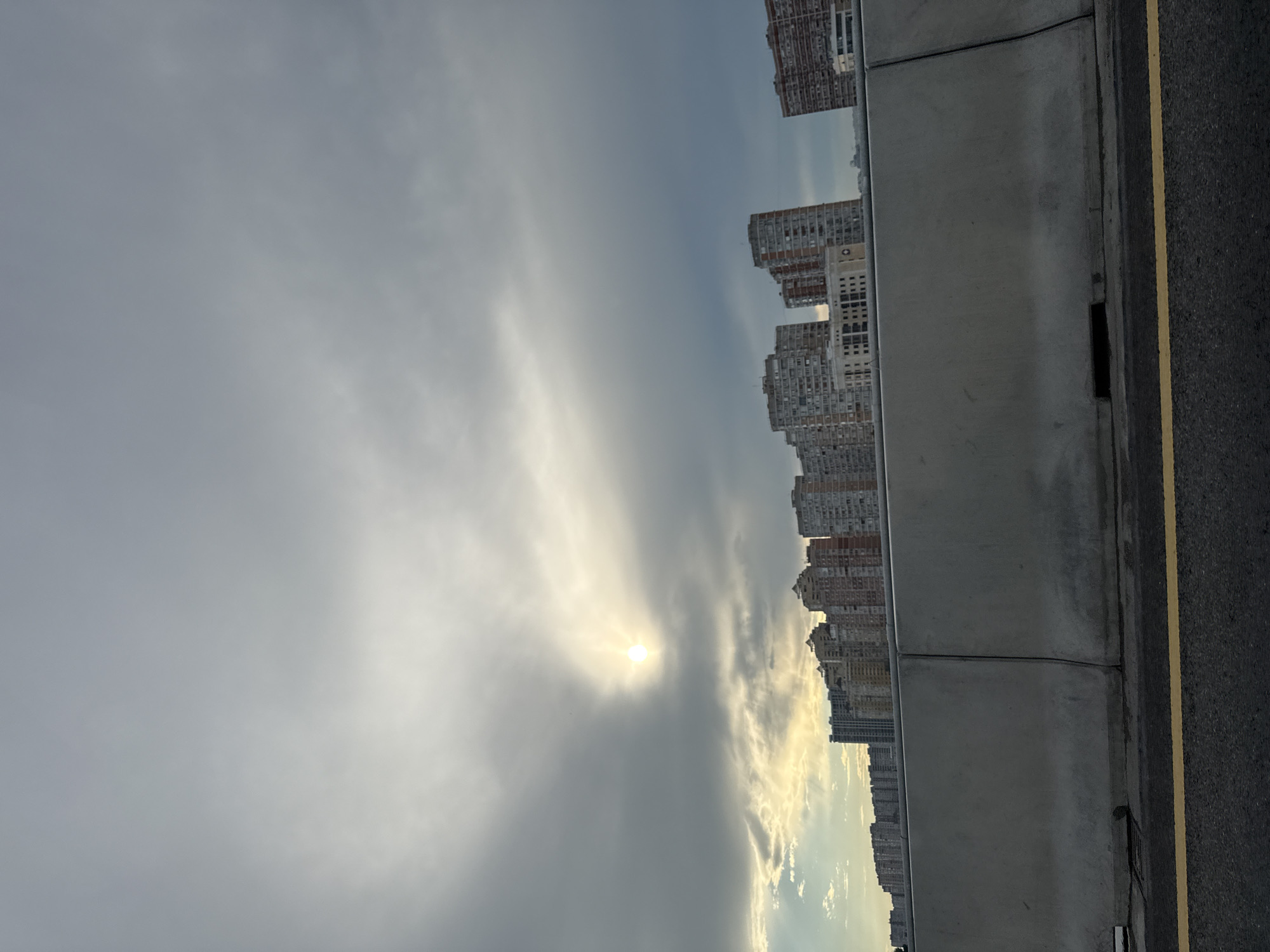 A wide shot captures a city skyline under a cloudy sky, with the sun peeking through the clouds. In the foreground, a concrete barrier runs horizontally across the frame, separating the road from the urban landscape. The barrier is textured and appears to be made of two joined sections. A bright yellow line runs along the edge of the asphalt road, which is visible at the bottom of the image. The city skyline features numerous apartment buildings of varying heights and architectural styles, some with visible windows and balconies. The sky is filled with grey and white clouds, with a luminous circle indicating the sun, casting a soft glow through a break in the clouds. The overall scene suggests an overcast day in an urban environment.