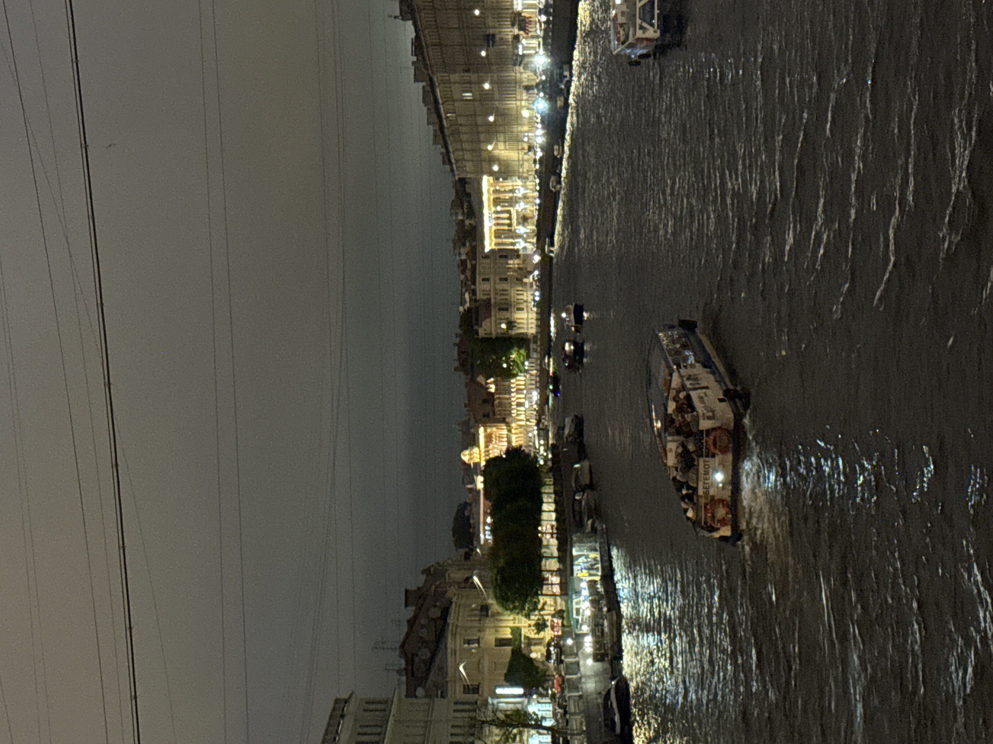 This is a nighttime photograph taken from an elevated vantage point overlooking a canal in Saint Petersburg, Russia. The canal is filled with dark, rippling water reflecting the lights from the surrounding buildings and boats. Several boats are visible on the canal. In the foreground, a tour boat, possibly a river cruise vessel, is moving from left to right, illuminated from within and by lights on its exterior. It appears to be carrying passengers. Further into the scene, other smaller boats and barges are docked or moving along the canal.
The canal banks are lined with multi-story buildings, some of which are grand and elaborately lit, displaying classic European architecture with many windows. Some buildings appear to be commercial establishments or historical landmarks. Trees are planted along the canal edges, and their foliage is dark and somewhat indistinct in the low light. The sky above is dark and overcast, with visible power lines or utility wires stretching across the frame horizontally. The overall atmosphere is one of urban nightlife, with the artificial lights contrasting with the dark sky and water. The specific location is likely one of the many canals that crisscross the historic center of Saint Petersburg, such as the Griboyedov Canal or the Moyka River, given the architectural style and the presence of tour boats.