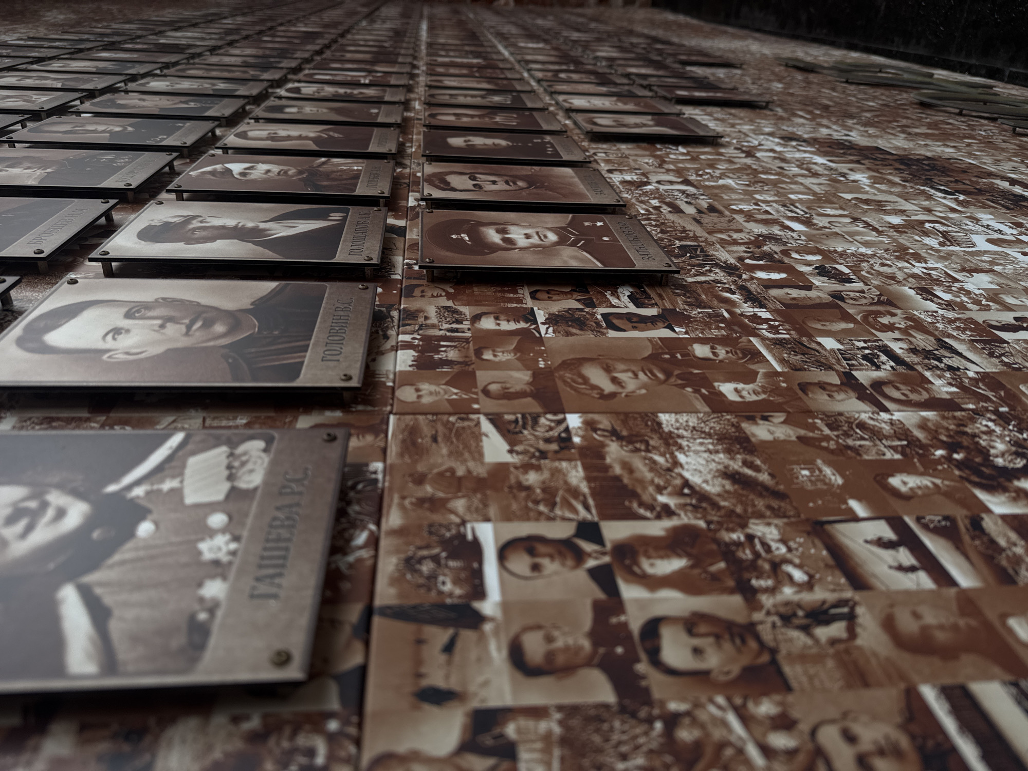 The image shows a memorial wall in Nizhny Novgorod, Russia. The wall is covered in sepia-toned photographs of people, both individually mounted and arranged in a larger mosaic. The individually mounted photographs are rectangular with metal borders, each containing a portrait and a name written in Cyrillic. The names visible include "ГАШЕВА Р.С.", "ГОЛОВИН В.С.", and "ГОЛОЩАПОВ А.К.". The background behind these mounted images is filled with smaller, tiled photographs creating a mosaic pattern. The angle of the shot is low and looking upwards, giving a sense of the wall's height and vastness.