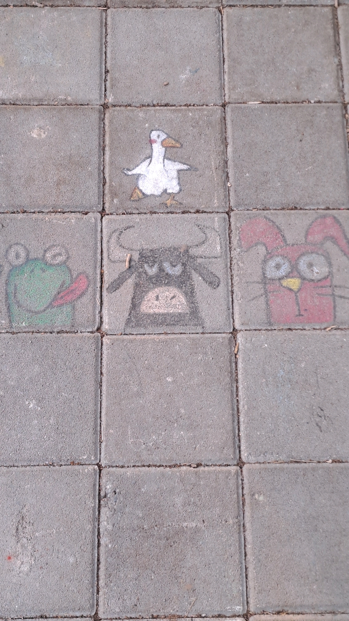 The image shows a close-up, eye-level view of a section of paved ground, likely a sidewalk or plaza. The ground is composed of square concrete pavers. Three of these pavers, arranged in a horizontal line in the center of the frame, feature chalk drawings of cartoon animals. On the left, a green frog with a pink tongue is depicted. In the center, a black bull with horns and an angry expression is drawn. To the right of the bull, a pink rabbit with large, googly eyes and long ears is illustrated. Above the frog and bull, another paver displays a drawing of a white duck. The remaining pavers are bare concrete, with some debris, such as small twigs and leaves, visible on the surface. The lighting suggests it is daytime, with diffused light, indicating an overcast sky or shaded area.