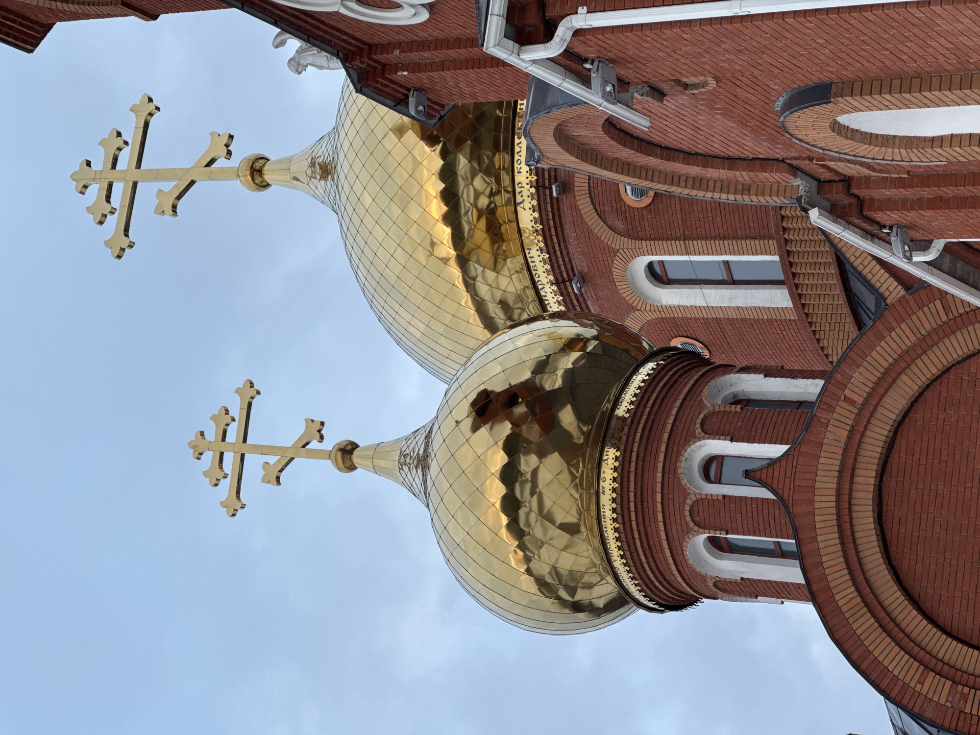 The image displays an upward-angled, close-up view of two golden, onion-shaped domes and a section of a red brick Orthodox church building in Chaikovsky, Russia, set against an overcast or light blue sky.
The two prominent domes are made of reflective golden material with a diamond-patterned surface. Each dome is surmounted by an intricate golden Orthodox cross, characterized by a triple horizontal bar and a slanted lower bar. The cross on the right-hand dome appears slightly larger and is positioned further back. The reflective surfaces of the domes show distorted reflections of the sky and surrounding architectural elements.
The church structure is constructed primarily from reddish-brown bricks, laid in various patterns, including arched brickwork above windows. Multiple arched windows with white frames are visible on the dome's supporting drum and the adjacent wall section. White rain gutters and downspouts are affixed to the right side of the brick wall, along with small white security or light fixtures. Cyrillic text, partially legible as "УПРАВЛЕНИЯ № 6" and "Дар коллектив," is inscribed in gold around the base of the larger dome. A small, indistinct white sculptural element, possibly an angel, is partially visible near the base of the rightmost cross. No individuals, actions, or interactions are depicted within the frame.