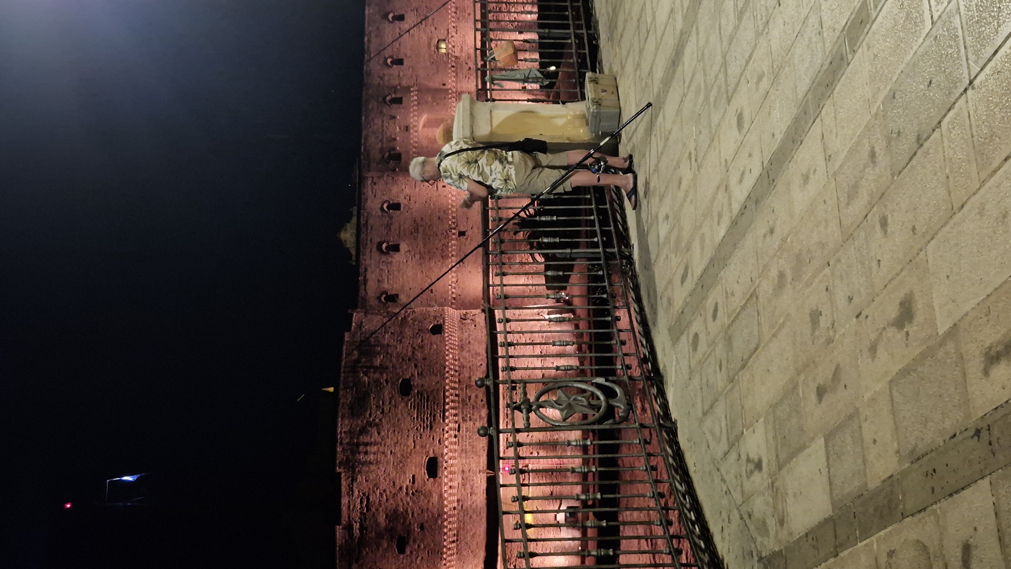 In Taranto, Italy, a man fishes at night by a castle. He stands on a paved walkway, holding two fishing rods. A red-lit building, likely a castle, is in the background. A metal fence with decorative elements separates the walkway from the water.