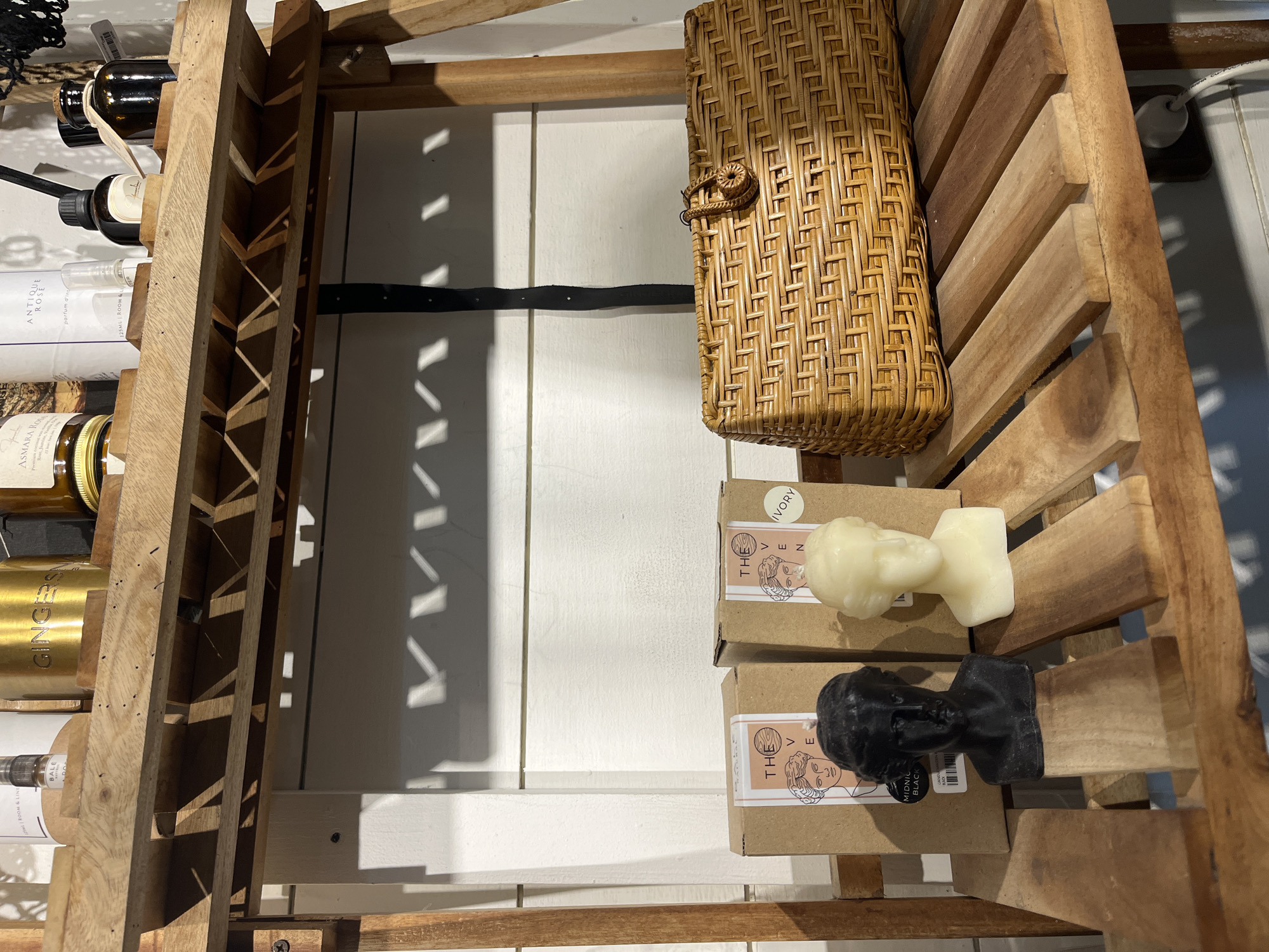 This image depicts a close-up, eye-level shot of a wooden shelf displaying decorative items. On the lower shelf, two bust-shaped candles, one black and one off-white, are positioned in front of two cardboard boxes with similar illustrations of faces. To the right of the candles, a woven wicker basket with a rounded handle is placed on the same shelf. The shelf itself is constructed of light-colored wood and features horizontal slats with gaps. Above this shelf, another wooden shelf is visible, holding various bottles and containers. The background consists of a white, horizontal planked wall with shadows cast upon it. The scene appears to be set indoors, likely in a retail or home decor environment. Based on its contents, this scene could be located in North Kuta, Indonesia, a region known for its artisan crafts and home goods.