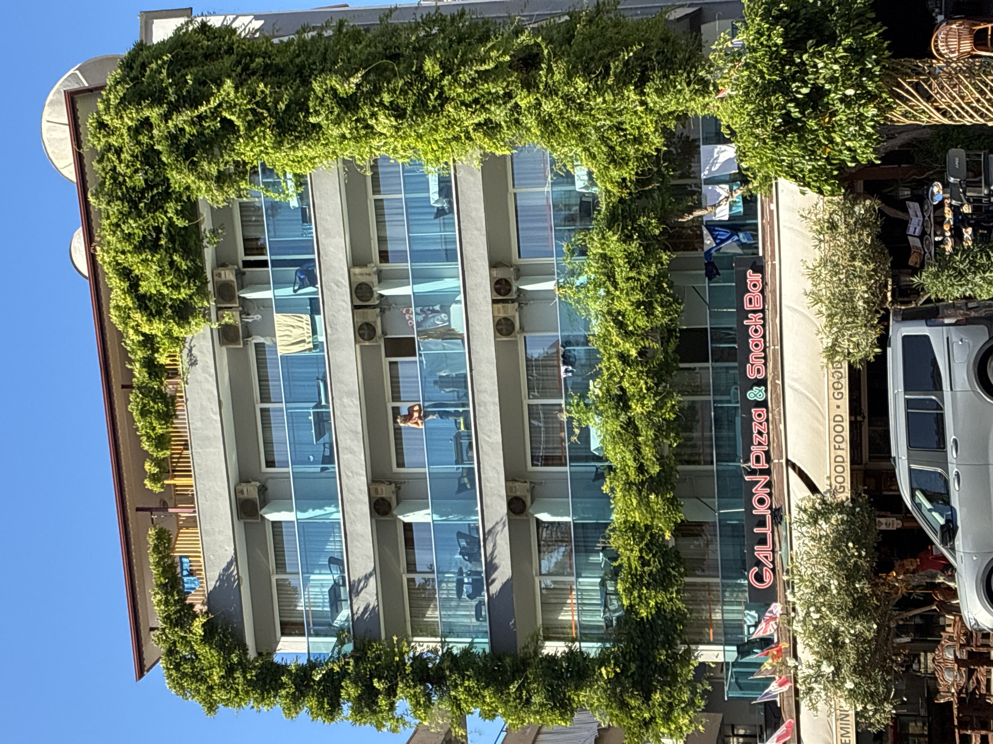 The image depicts the exterior of a multi-story building in Alanya, Turkey, prominently featuring a facade adorned with lush green vines. The building has several floors with balconies equipped with blue glass railings. On one of the balconies, a person is visible standing and looking outwards. Air conditioning units are mounted on the exterior walls. Below the balconies, a sign reads "GALLION Pizza & Snack Bar" in neon lettering. Several flags are displayed in front of the establishment. In the foreground, a silver van is parked, and outdoor seating with tables and chairs is visible, along with some greenery and trees. The sky is clear and blue, indicating a sunny day.