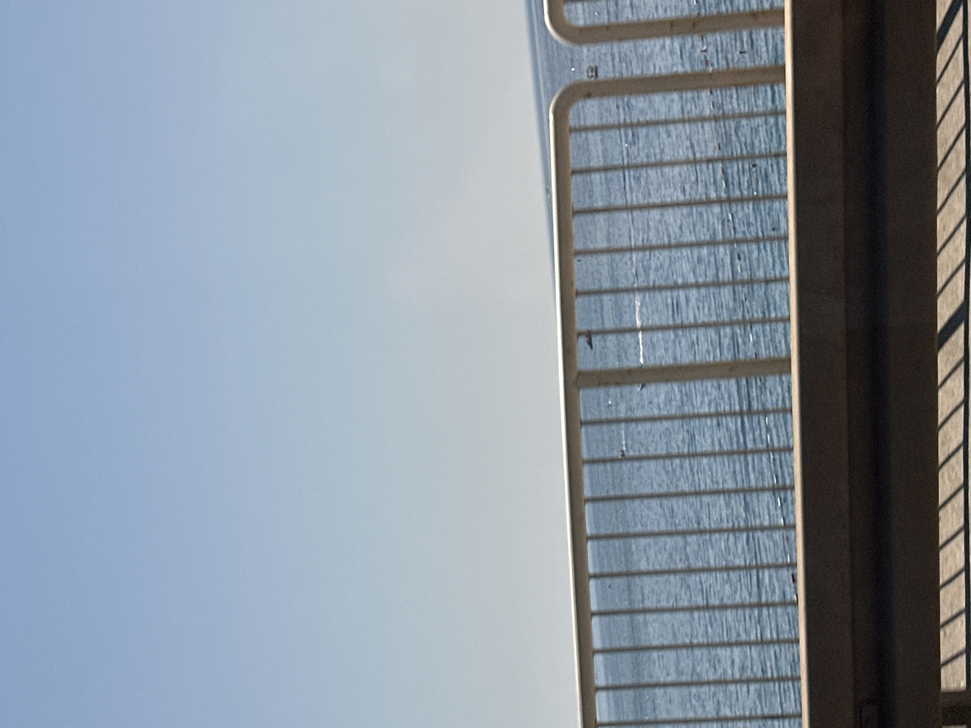The image depicts a clear blue sky above a body of water. In the foreground, a metal railing with vertical bars separates the viewer from the water. The water appears to be calm with small ripples. There are a few dark objects scattered on the surface of the water, some of which could be small boats or buoys. A concrete structure is visible below the railing, and shadows are cast on a surface beneath it, suggesting the presence of a walkway or road. The scene is consistent with a view from a promenade or bridge overlooking the sea in a coastal city. Given the information about Budva, Montenegro, this could be a view of the Adriatic Sea from a promenade or a bridge along the coast.
