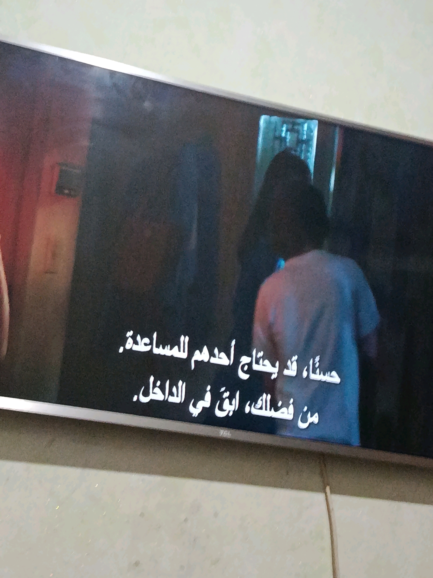 The image is a close-up, slightly angled shot of a television screen displaying subtitles in Arabic. The subtitles read: "حسناً، قد يحتاج أحدهم للمساعدة، من فضلك، ابق في الداخل." which translates to "Okay, someone might need help, please, stay inside." The screen content is the primary focus, with a dark, grainy image of what appears to be a scene from a movie or show playing on the television. There is a person visible from the back, wearing a light-colored t-shirt, in the right half of the screen. The background on the screen is dark and blurry, with some indistinct shapes and colors. The television itself has a thin bezel and is mounted on a wall. The wall texture is visible above and below the screen. There is no discernible location in Kenitra, Morocco, as the image only shows a television screen and its immediate surroundings.
