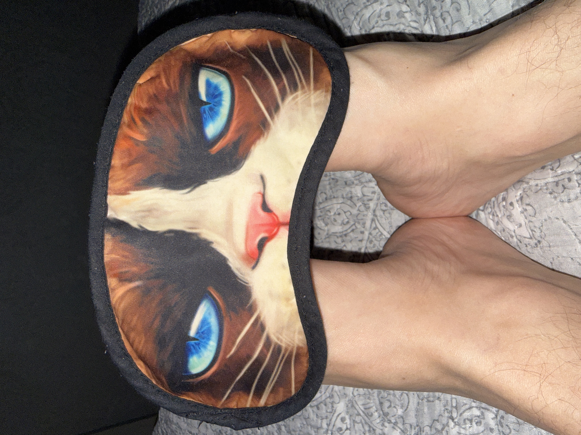 The image shows a pair of bare feet resting on a patterned gray surface. A cat-themed sleep mask is positioned between the feet. The feet are light-skinned, and the mask features a cat face with blue eyes. The surroundings appear to be an indoor setting. There is no location specified within the image.
