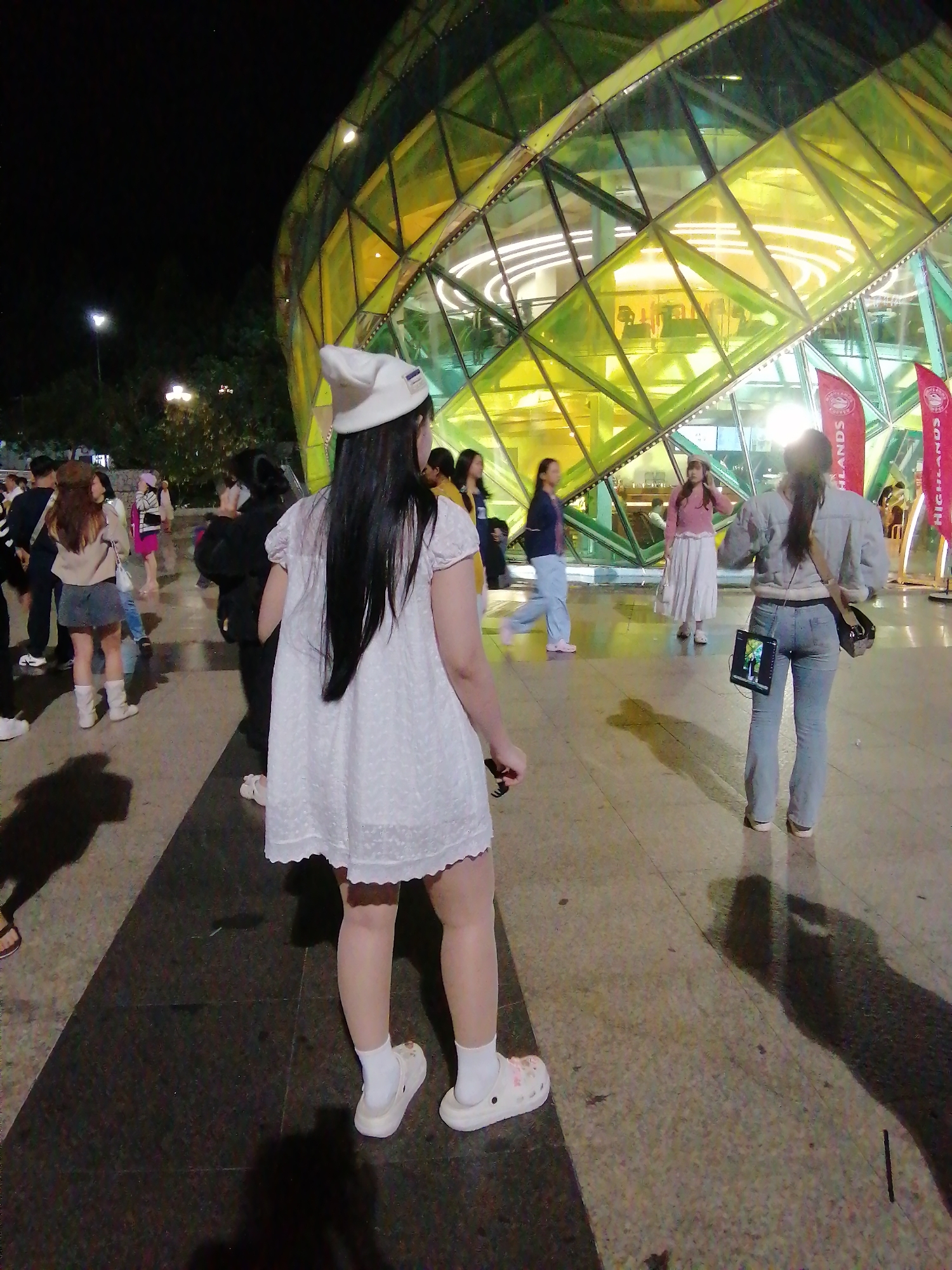The image captures a group of people gathered outdoors at night, in front of a strikingly illuminated, geodesic dome-like structure with a faceted glass facade in shades of green and yellow. The building appears to be a commercial or entertainment venue, possibly a shopping mall or a concert hall, given the presence of red banner-like flags with text.
In the foreground, a young woman with long, dark hair, wearing a white beanie, a short white dress, white socks, and white crocs, is the central subject. She is seen from behind, looking towards the building. Other individuals are dispersed around the area, some walking, some standing and conversing. A woman in a denim jacket and jeans is visible further back, with a tablet attached to her back. Another woman in a pink top and a long skirt is also present.
The scene suggests a lively evening gathering or event. The lighting from the building and some streetlights illuminates the area, casting shadows on the ground, indicating it's nighttime. The overall atmosphere seems relaxed and casual, with people enjoying the urban nightscape. The presence of flags with text, though partially obscured, might indicate a specific event or brand associated with the location. The location context of Da Lat, Vietnam, doesn't provide specific visual cues in this particular frame, but the architectural style and the people's attire are consistent with a modern urban setting.