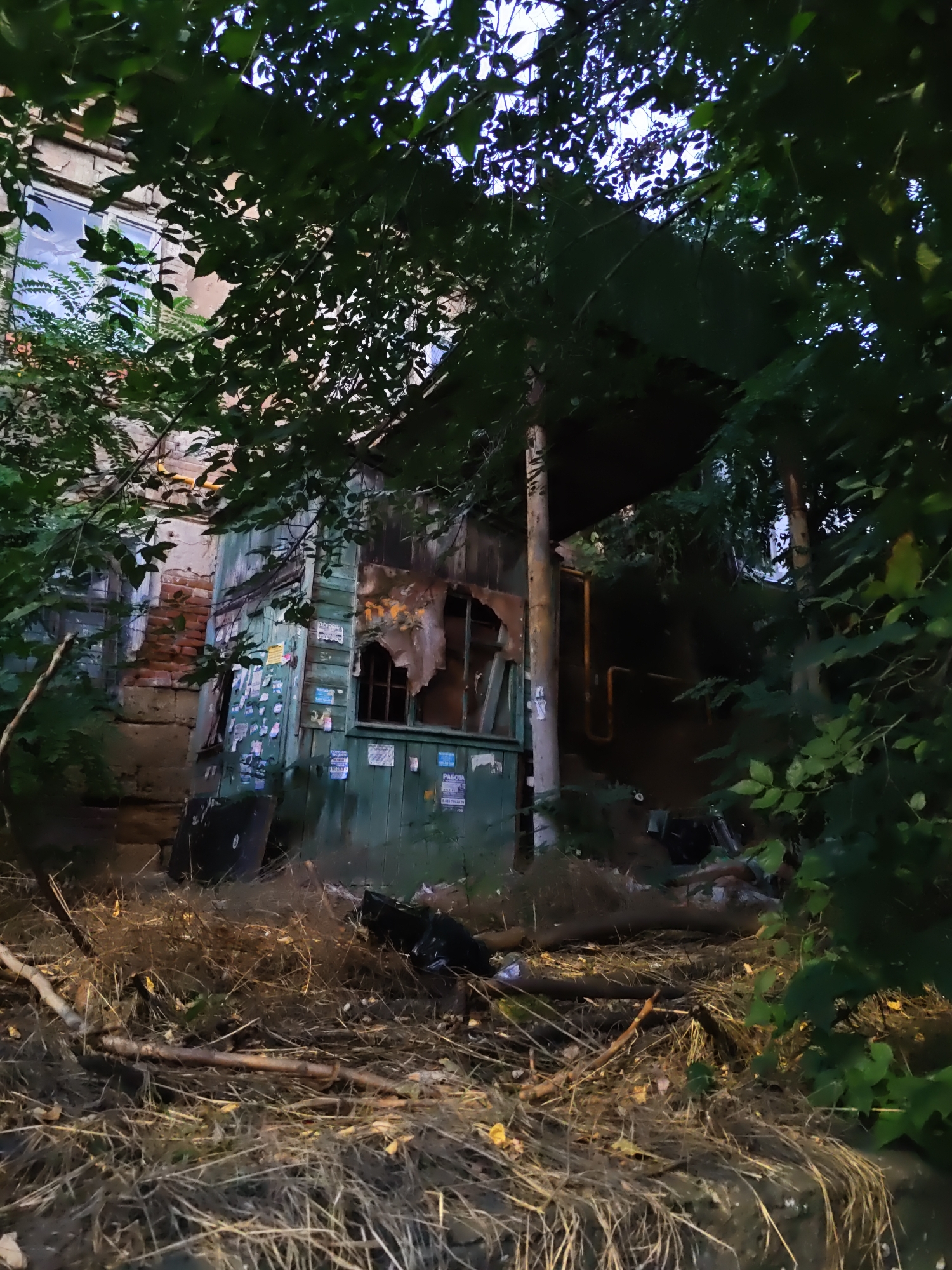 This image depicts an abandoned, dilapidated structure with overgrown vegetation in Rostov-on-Don, Russia. The building appears to be constructed of wood and brick, with peeling paint and broken windows. Debris, including dry grass, branches, and what looks like trash bags, is scattered around the foreground. The overall impression is one of neglect and decay. The lighting suggests it is either dusk or early morning.