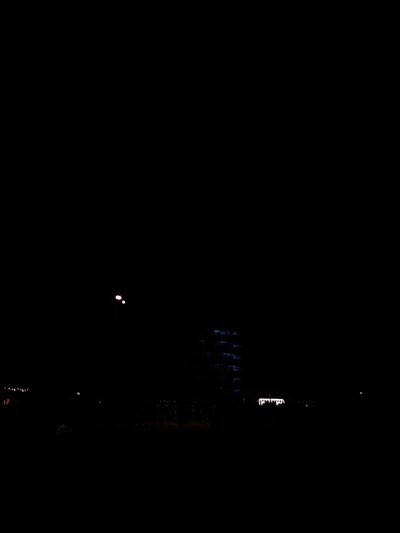 The image shows a night scene in Indio, United States. A tall building with illuminated windows is visible in the distance. There are streetlights and other lights lining a road or street. Some trees or bushes can be seen in the foreground.