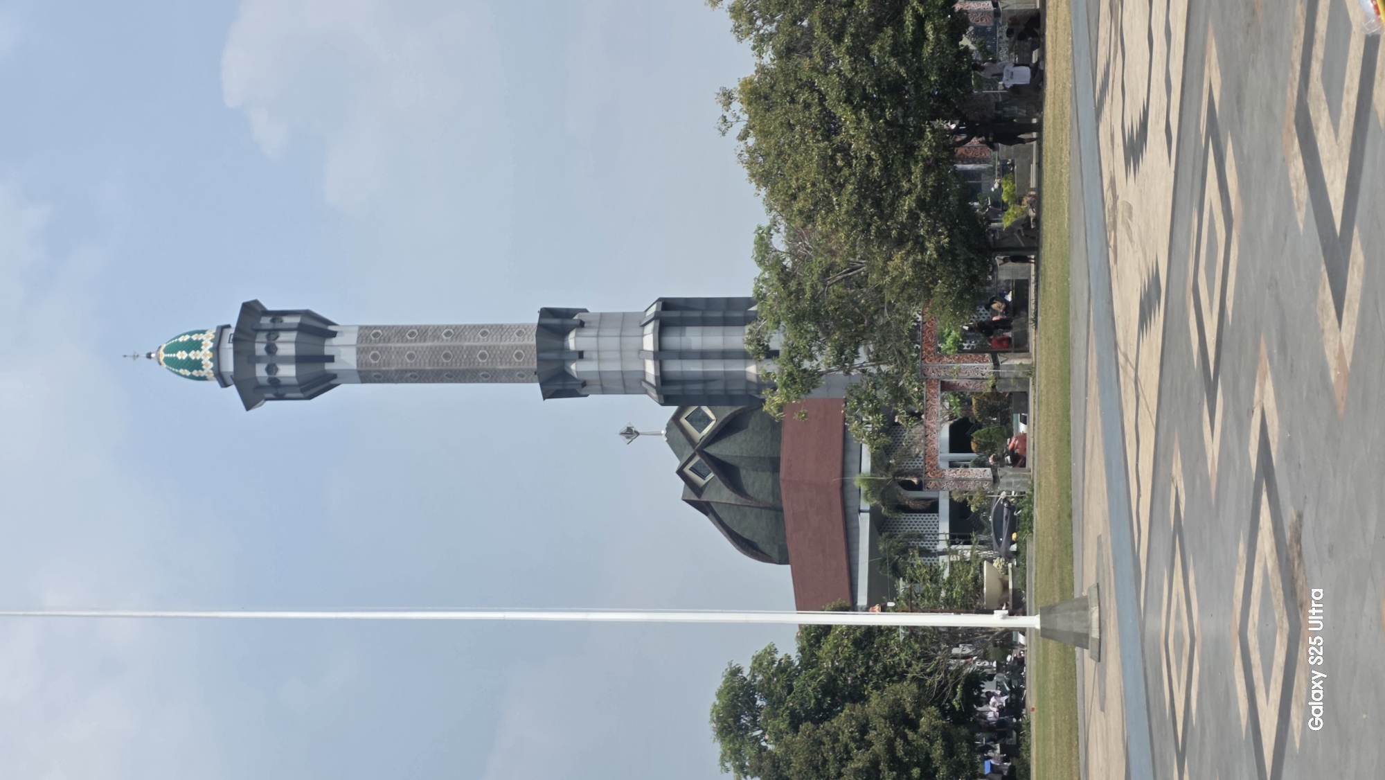 The image shows a mosque with a tall, slender minaret topped with a green dome. The minaret features a patterned shaft and several levels of balconies. Behind the minaret, a large, dark green dome with geometric patterns is visible, along with a red-tiled roof and part of the mosque's facade. The mosque is surrounded by lush green trees. In the foreground, there is a paved area with a geometric design, a grassy field, and a tall flagpole. Several people are scattered in the background, some sitting on benches or walking in the open space. The sky is partly cloudy. This scene is likely located in Kecamatan Pati, Indonesia, given the architectural style and common presence of mosques in the region.