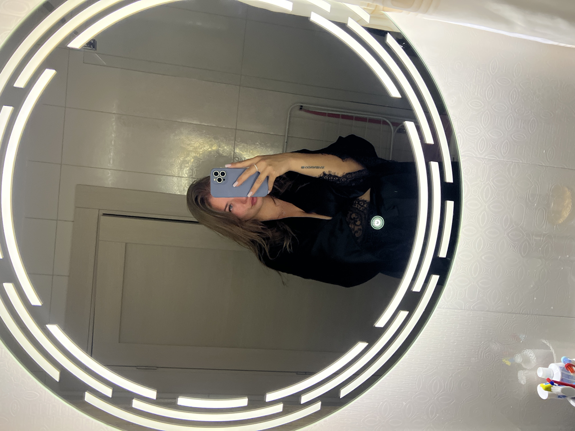 A young woman is taking a selfie in a bathroom mirror. The mirror is round and lit with LED lights. The bathroom has white tiled walls with a faint pattern. Toothbrushes and toothpaste are visible on a shelf or counter. The woman is wearing a black robe or top with lace detail. She is holding a blue smartphone. A tattoo is visible on her left arm. In the background, a white drying rack can be seen. It is not possible to confirm the location as Budva, Montenegro from the given image.