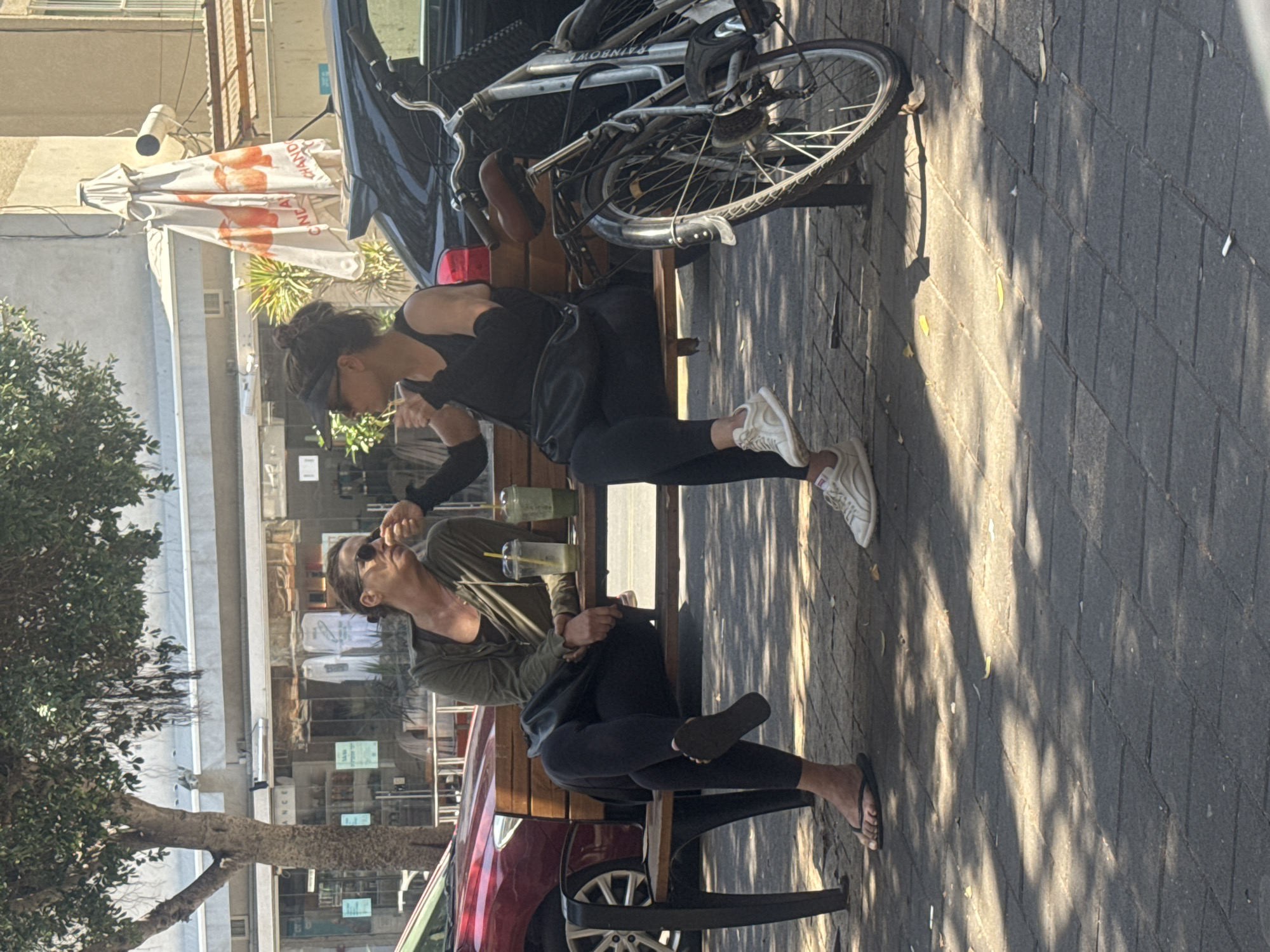 Two people sit on a public bench facing opposite directions. Both are drinking beverages with straws from clear cups. The person on the top of the image wears black clothing and white sneakers. The person on the bottom of the image wears a light green shirt and dark-colored leggings and footwear. A black car and a red car frame the bench. A bicycle rests against the back of the bench. The image appears to be taken outdoors in Tel-Aviv, Israel, evident from the architecture and signs visible in the background. The foreground shows a sidewalk with dappled sunlight.