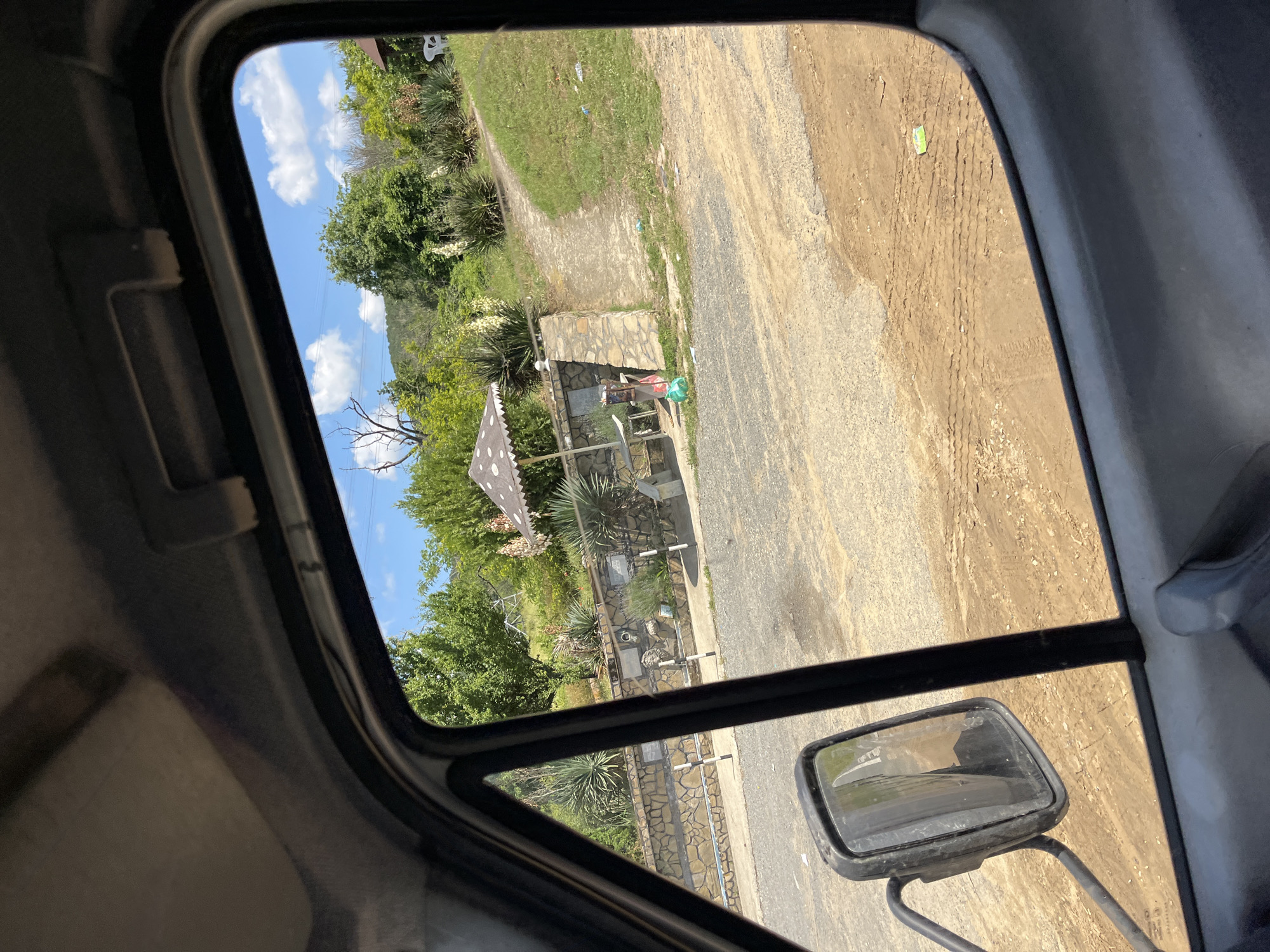 The image, taken from inside a vehicle looking out a window, depicts an outdoor scene in Prosenik, Bulgaria. The foreground shows the vehicle's interior with a portion of the dashboard and a side-view mirror. The window frames a view of a dusty, gravel road leading to a green, grassy area. In the middle ground, there's a stone wall with several decorative plaques and a lion head sculpture. A few plants, including agave-like succulents and tall flowering plants, are visible. An umbrella with a mushroom-like pattern shades a small table and chairs. Further back, lush green trees and hills form the background under a blue sky with scattered white clouds. Several pieces of litter are scattered on the ground in the grassy area.